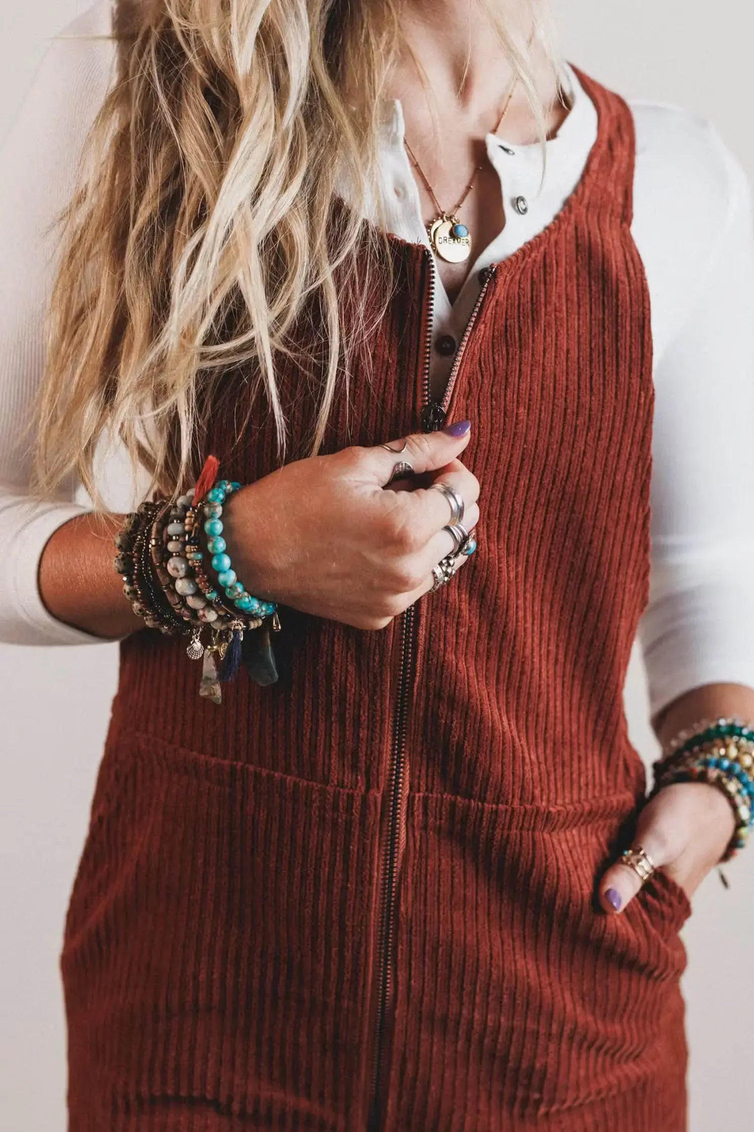 A close-up of a woman wearing a rust-brown corduroy zip-front vest over a white long-sleeve top, accessorized with multiple bracelets and rings.