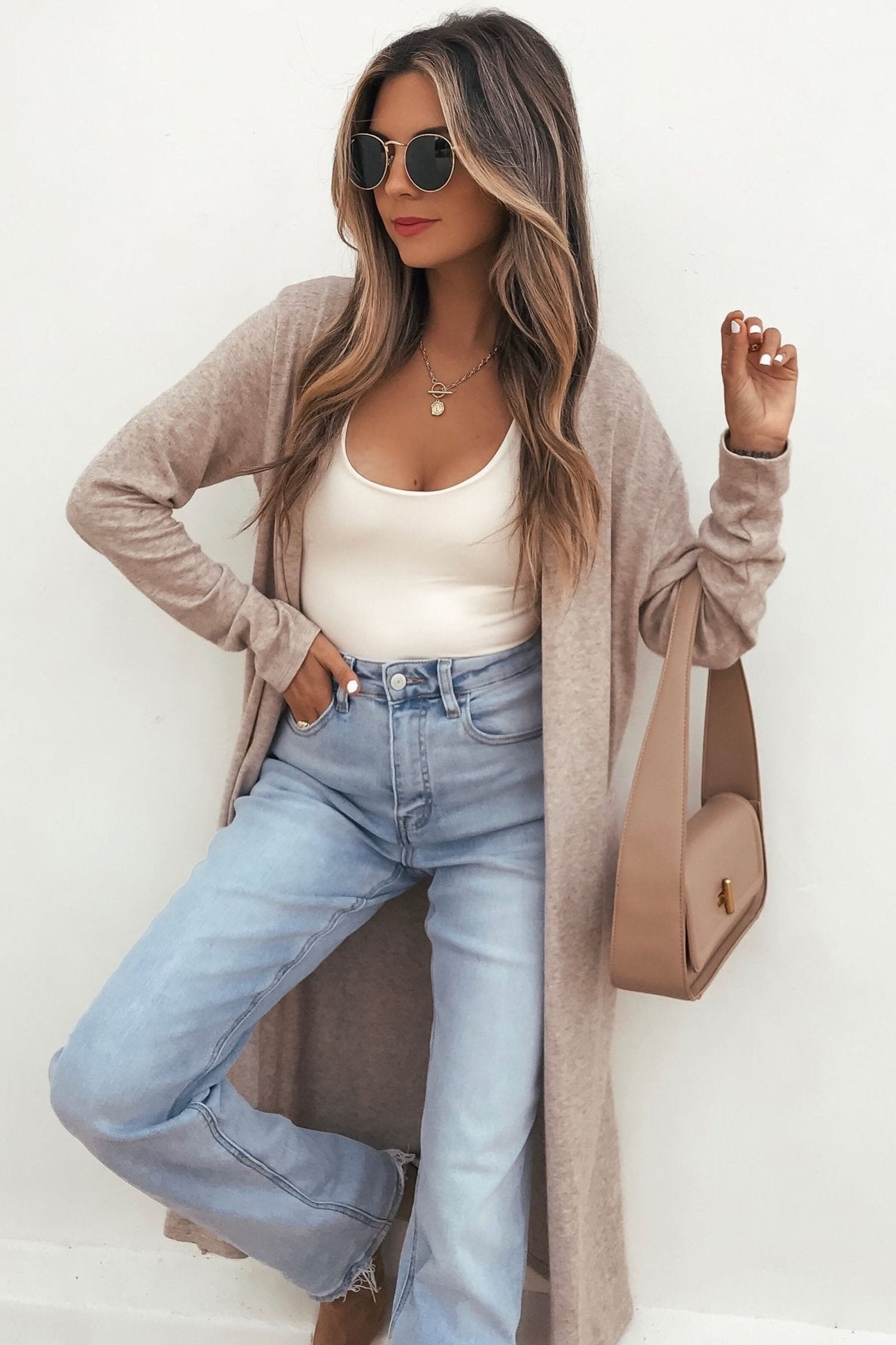 Woman in taupe knit cardigan, white tank, light blue jeans, and matching shoulder bag.