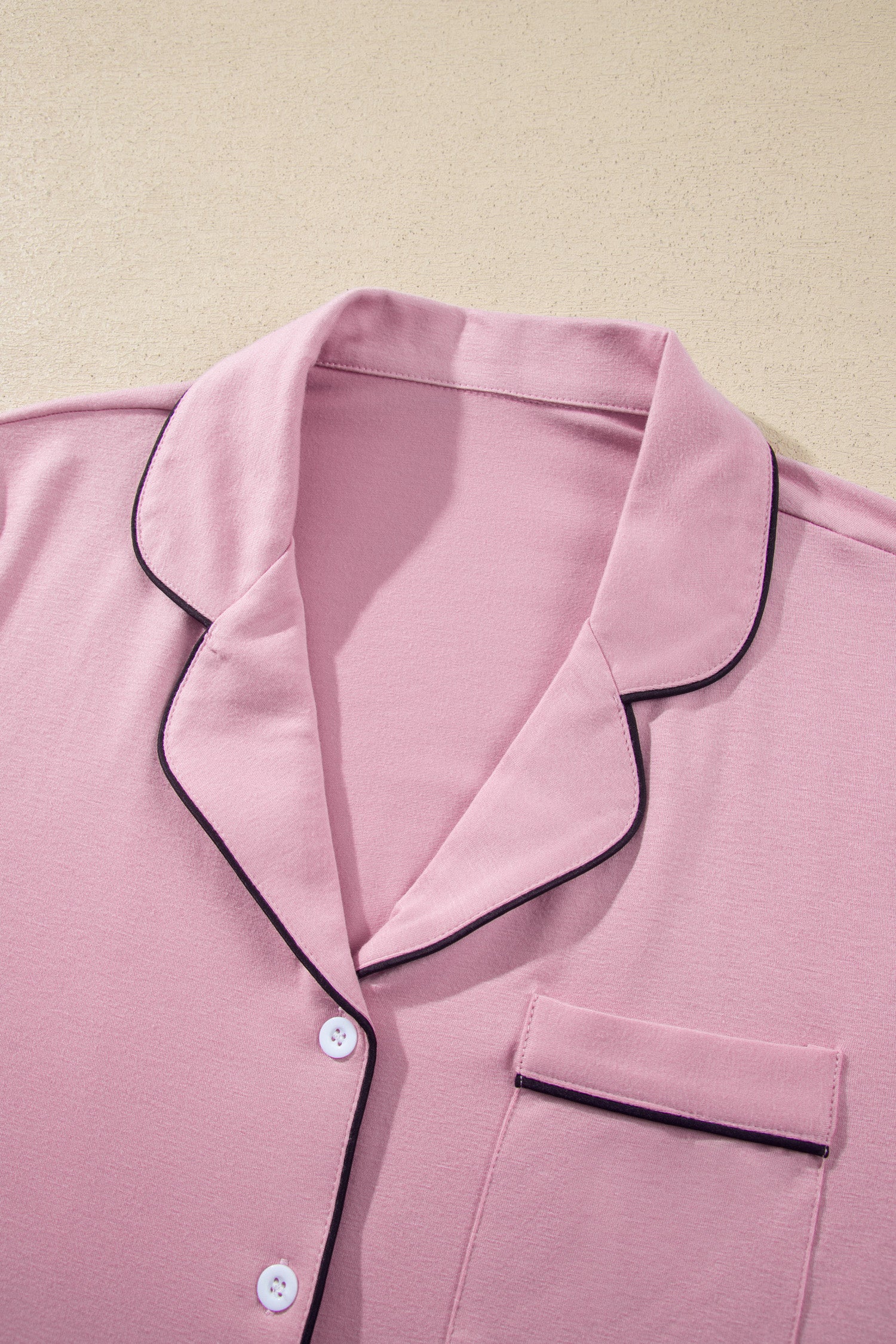 A close-up of a pink pajama top with black piping trim, featuring a collar, button placket, and chest pocket, laid flat against a light beige background.