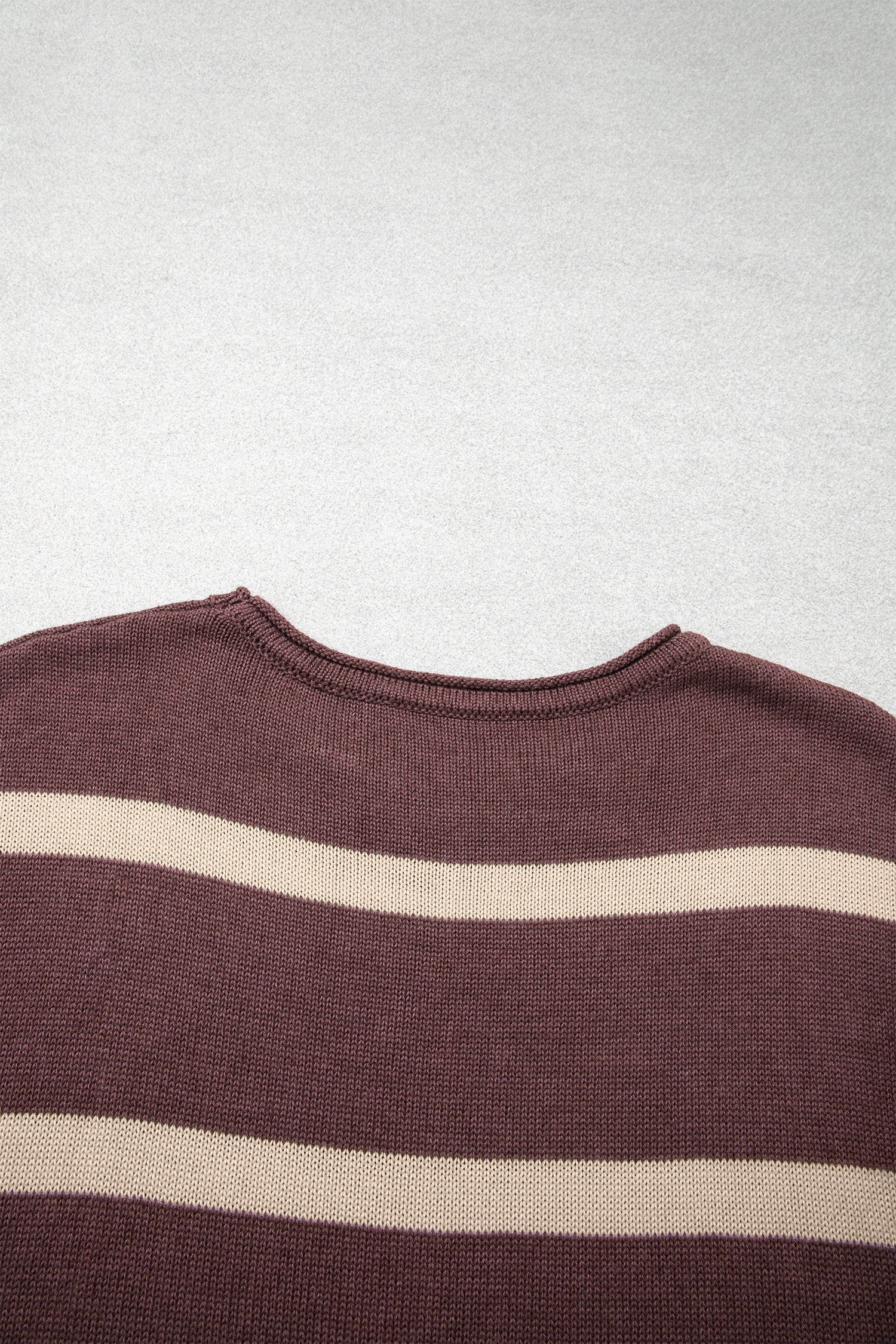 A close-up of a ribbed knit crewneck sweater with horizontal stripes in dark brown and cream, laid flat against a light gray textured background.