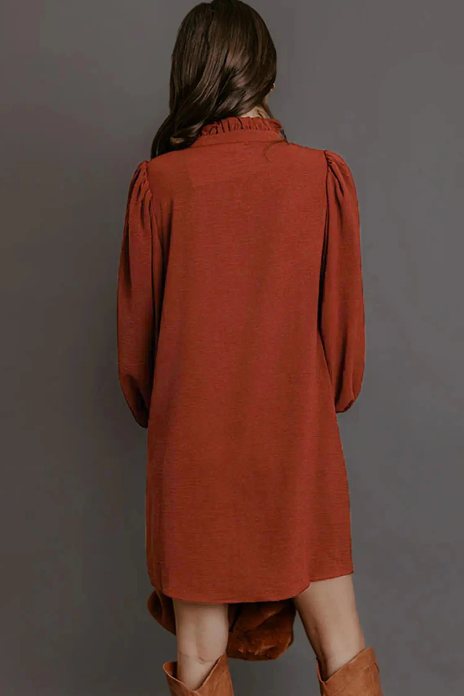 A woman viewed from behind wearing a rust-orange long-sleeve tunic dress with puffed sleeves and brown boots, set against a solid dark gray studio background.