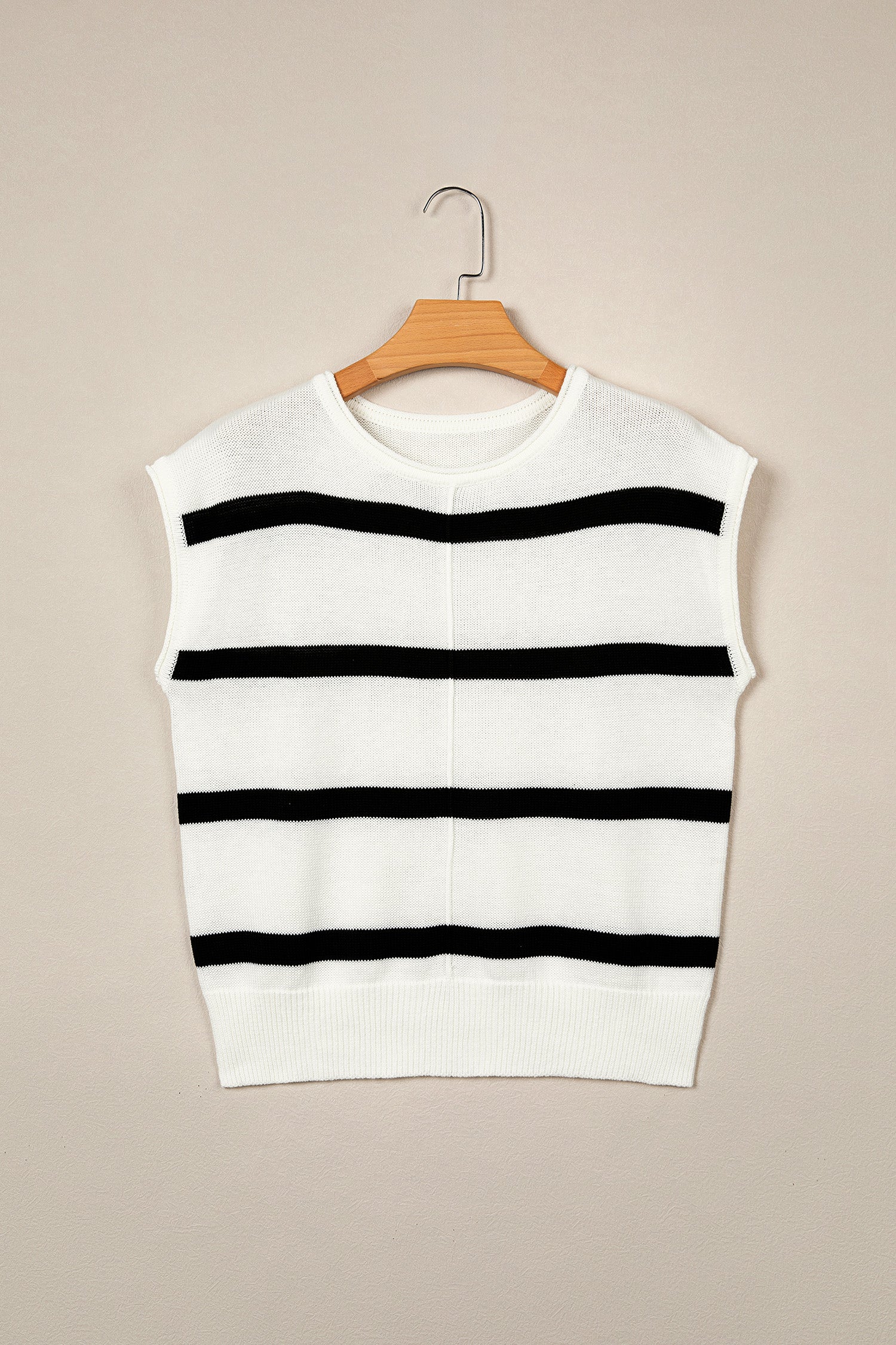 A sleeveless white knit sweater vest with horizontal black stripes, hanging on a wooden hanger against a solid beige background.