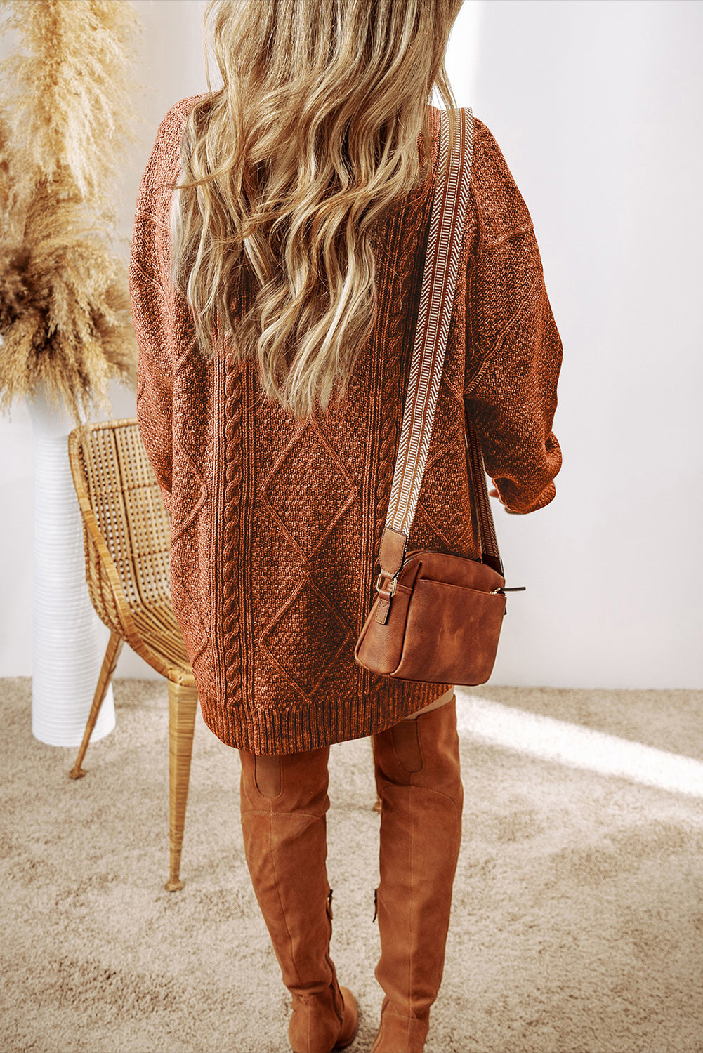Person wearing a brown knitted cardigan and over-the-knee boots, standing in a room with a chair and decorative items.