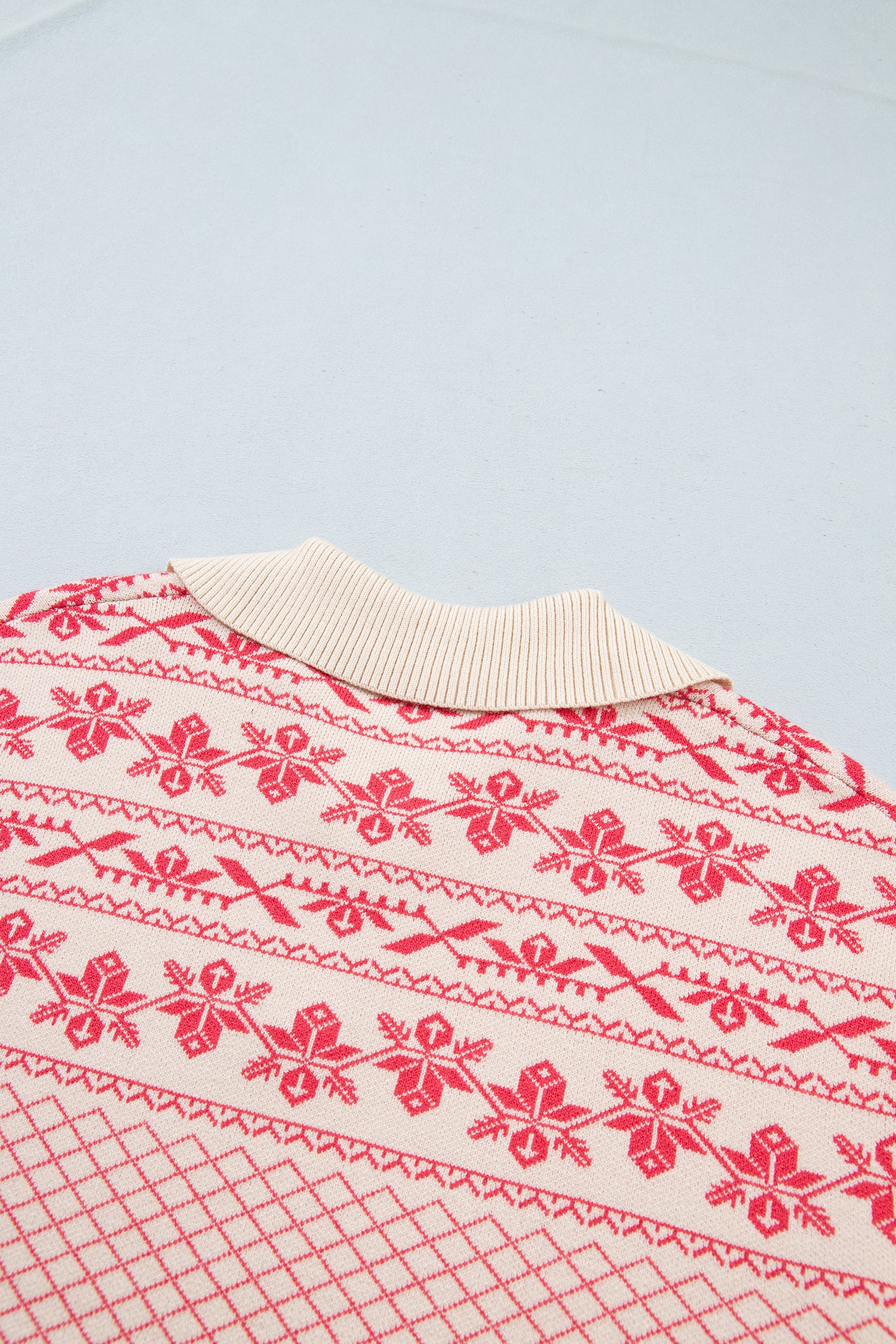 A close-up of a knit sweater featuring a red and cream Nordic pattern with a ribbed cream collar, laid flat on a light gray background.