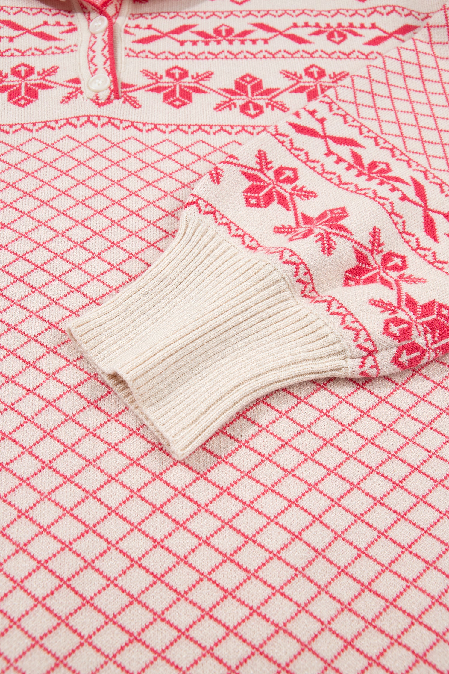 Close-up of a knit sweater with a cream base and red Nordic-style patterns, including a red diamond lattice and red snowflake motifs, with a ribbed cream cuff.