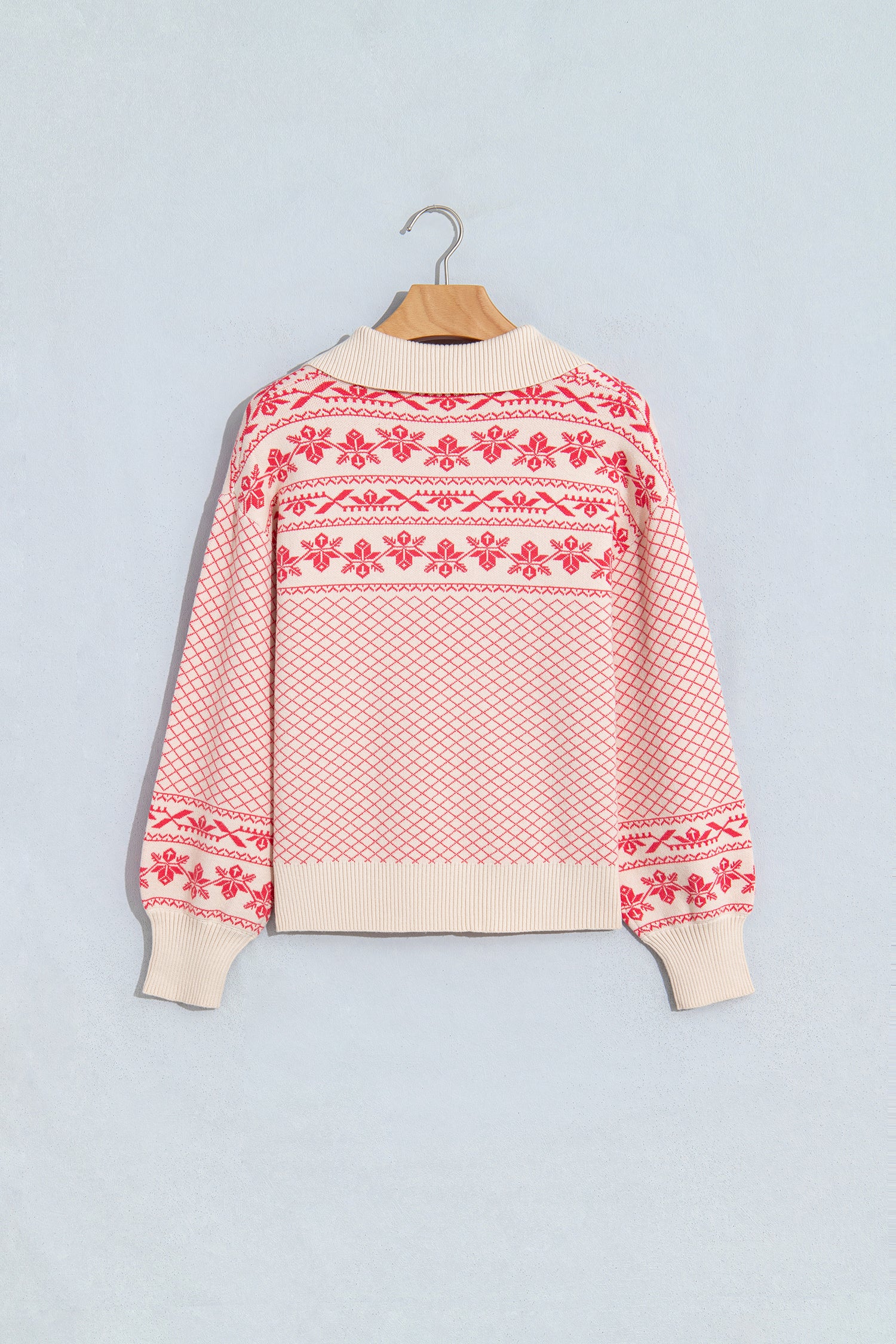 A cream-colored knit sweater with red Nordic and Fair Isle patterns, hanging on a wooden hanger against a light 