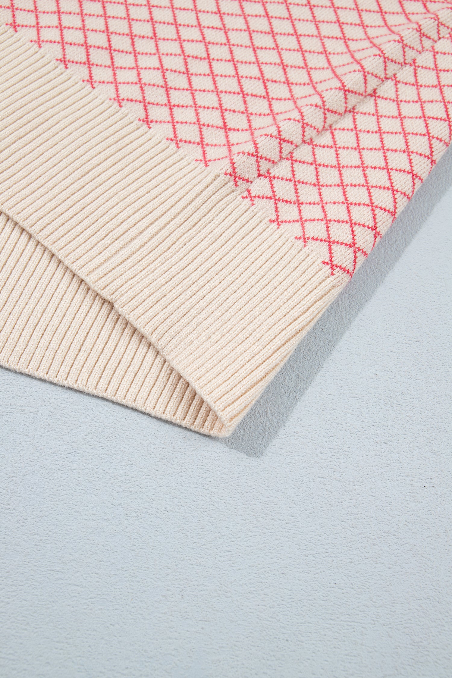 A close-up of a folded ribbed knit textile, showing a cream-colored ribbed border and a cream base with a red diamond grid pattern on a light gray background.