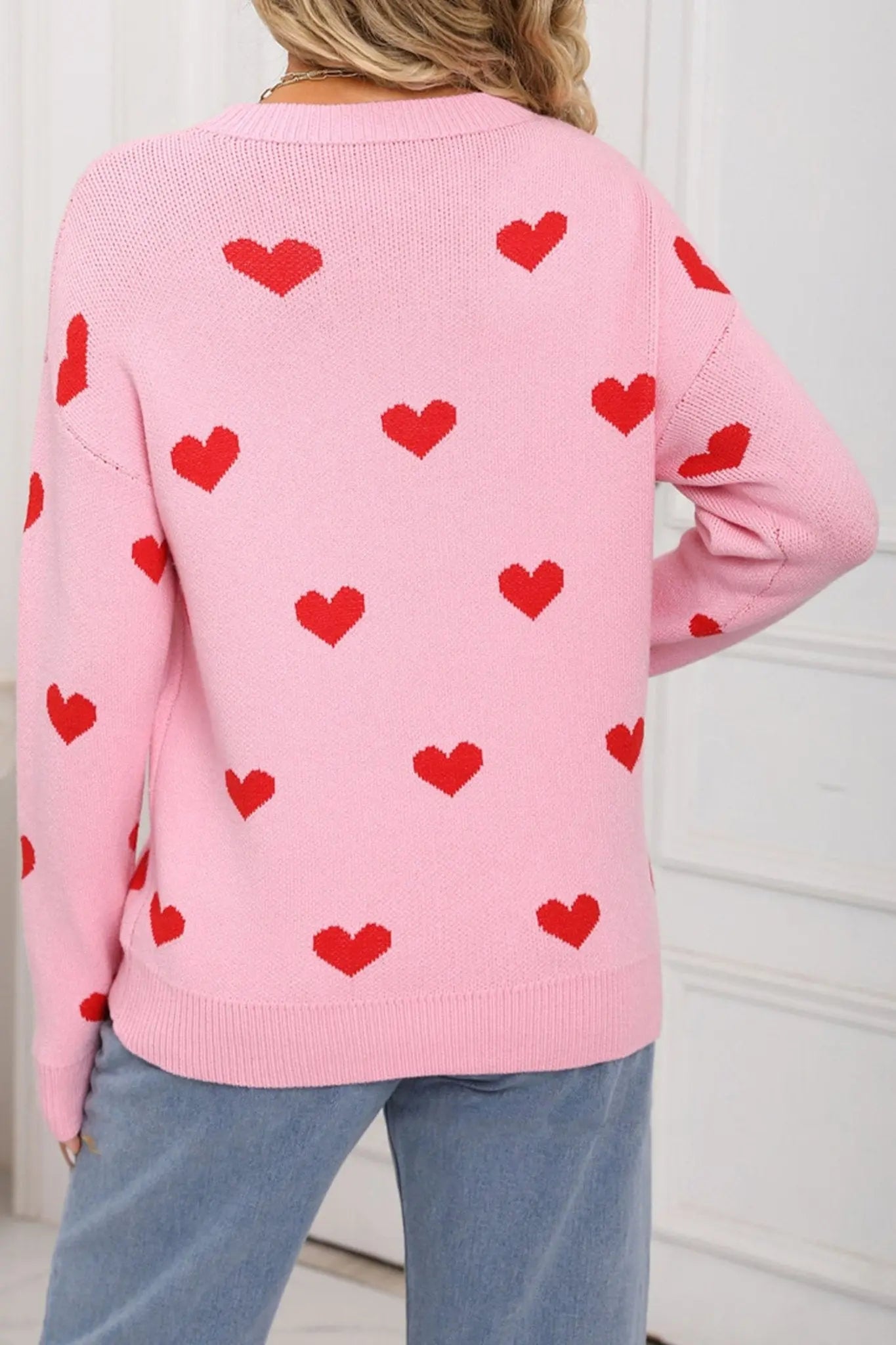 Pink sweater with red heart patterns worn by a person against a white background