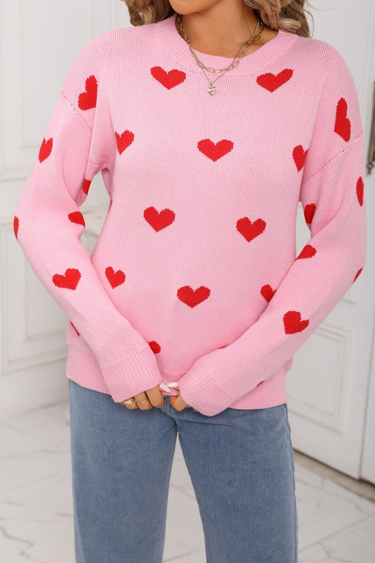 Person wearing a pink sweater with red heart patterns and blue jeans indoors.