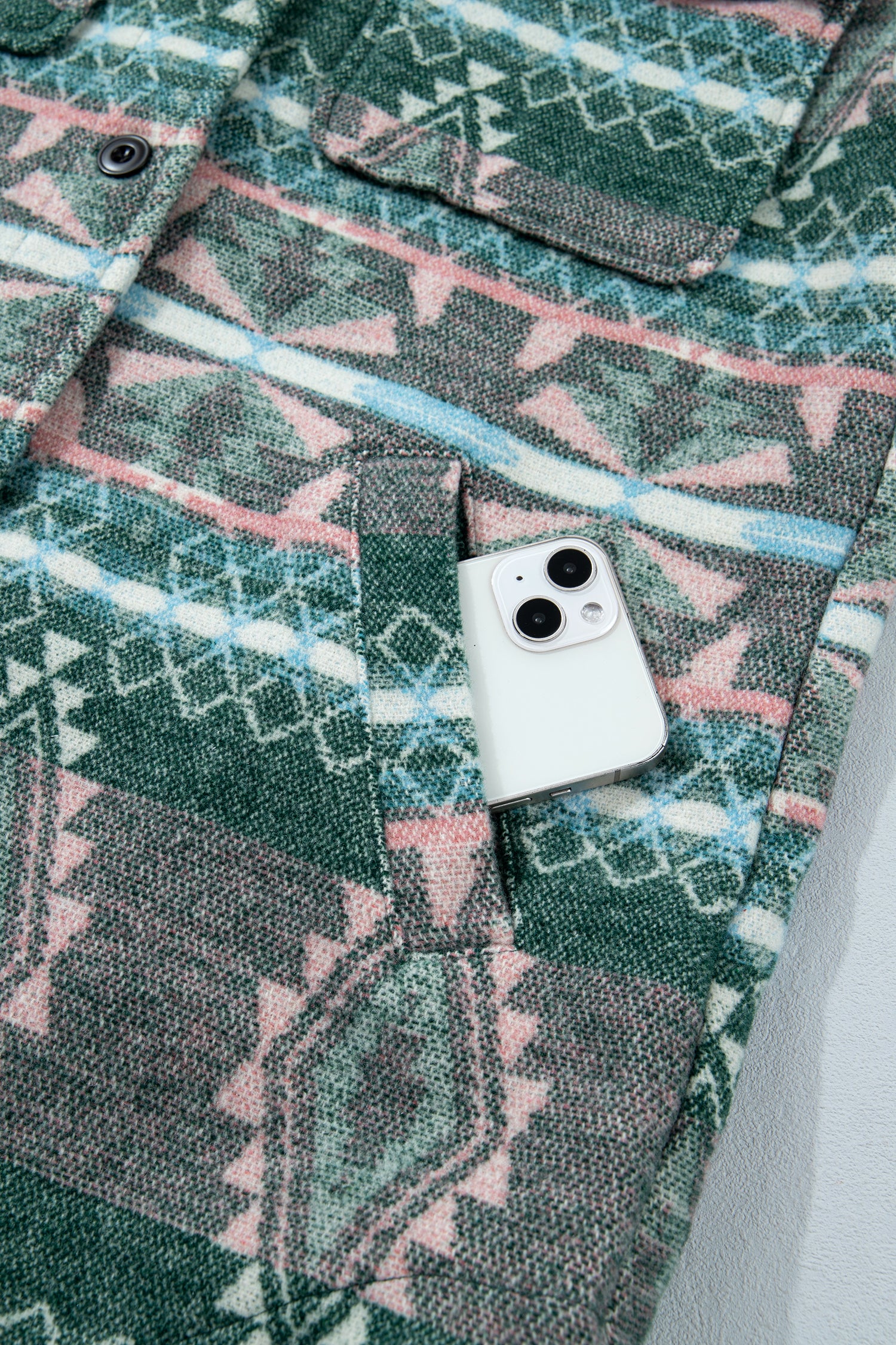 A close-up of a patterned pocket jacket or overshirt with a geometric Southwestern-style print in green, pink, and light blue. A white smartphone is placed inside one of the front pockets to demonstrate the pocket's capacity.