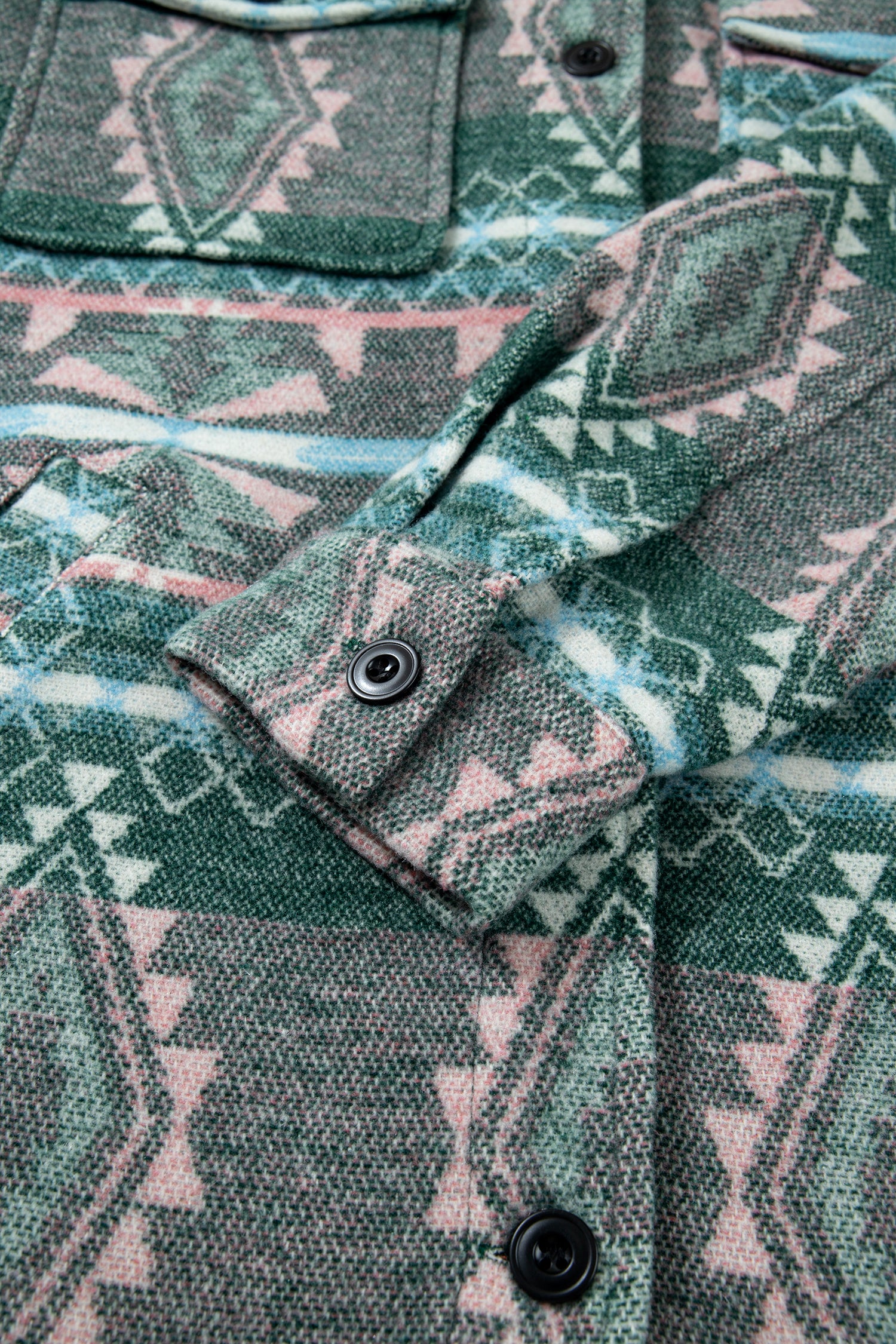 Close-up of a patterned button-up shirt or jacket fabric with a geometric Southwestern-style print in green, pink, and white, featuring a buttoned cuff and dark buttons.