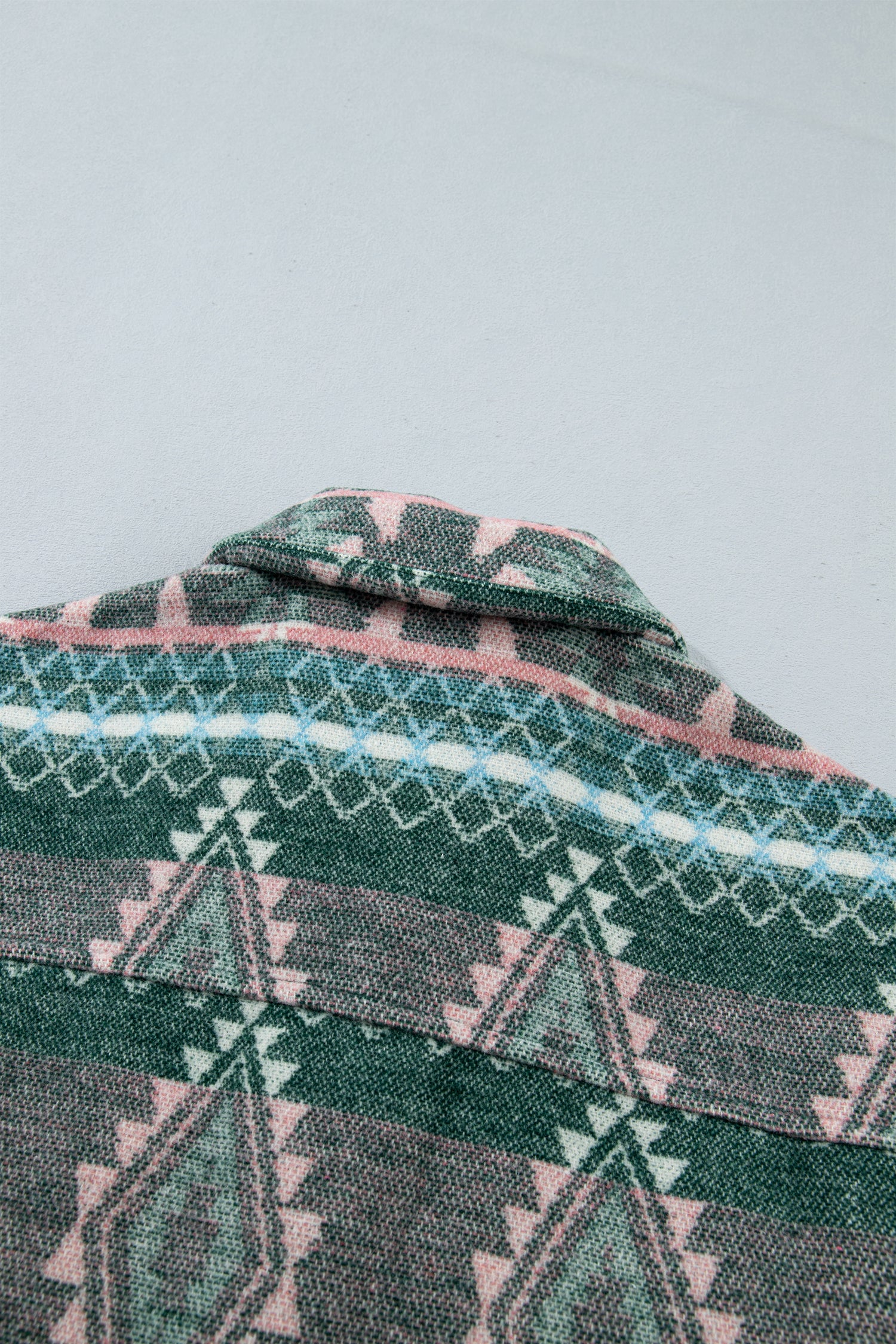 A close-up of a patterned woven textile, likely a shirt or blanket, featuring a geometric tribal design in dark green, pink, gray, and light blue.