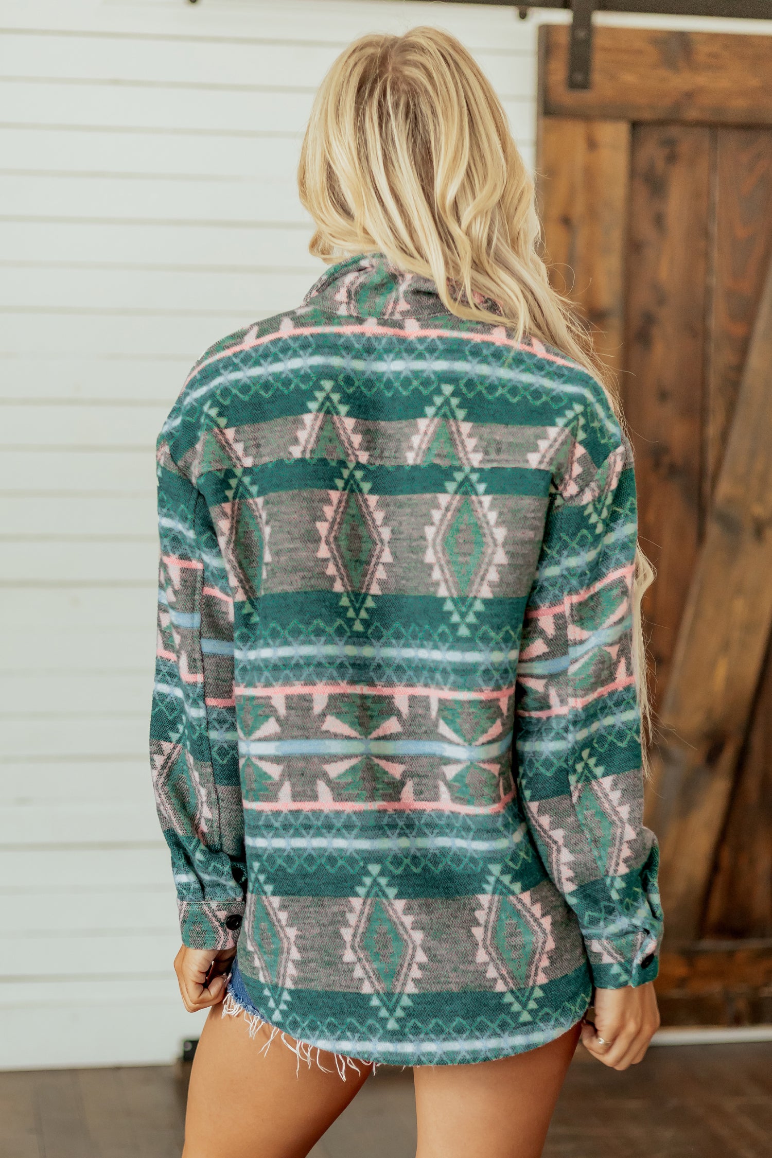 A woman viewed from behind wearing a long-sleeve patterned shirt with a green, gray, and pink geometric design, standing in front of a white siding and wooden door.