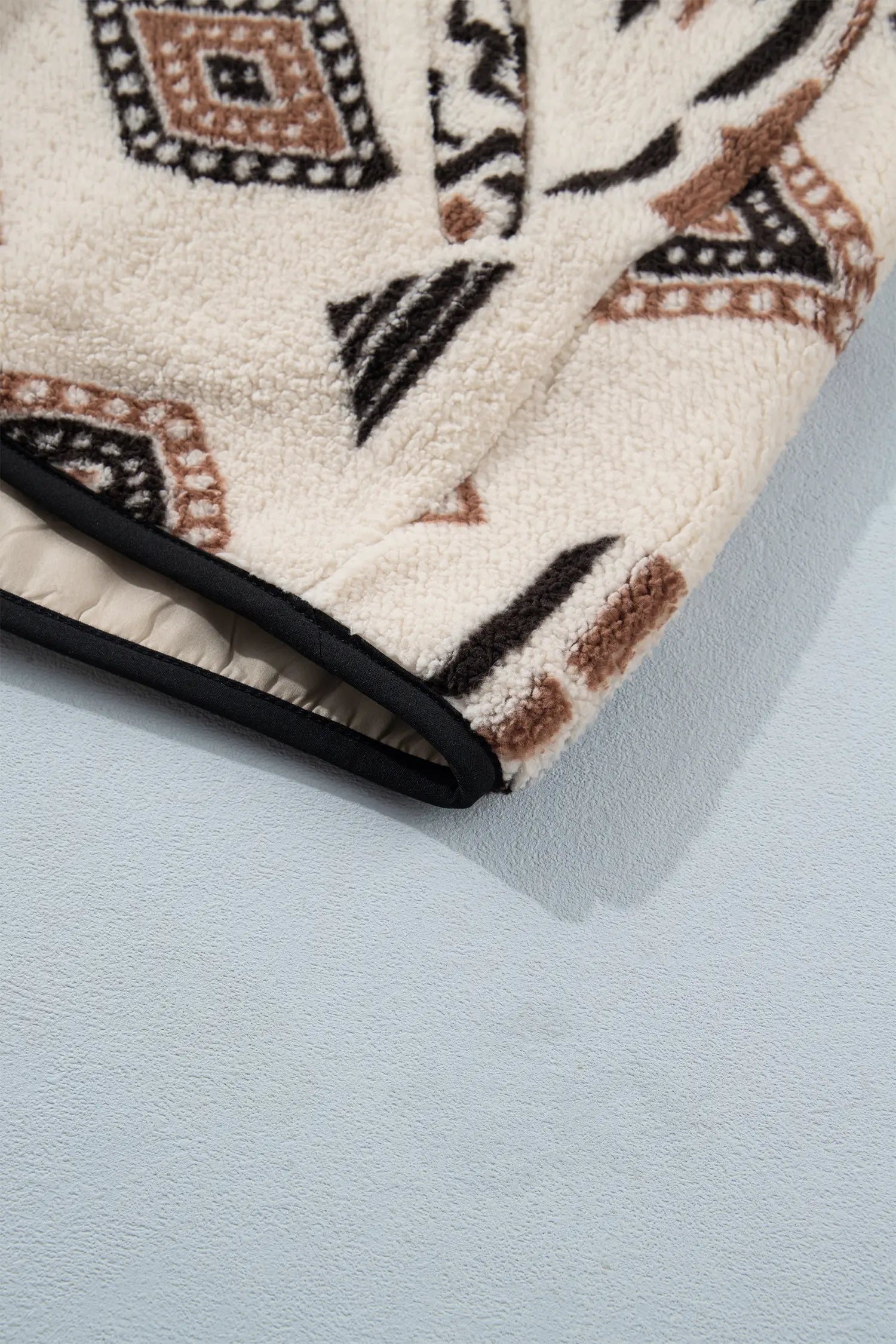 Close-up of a plush fleece blanket with a geometric pattern in black, brown, and beige, folded over to show its black stitched edge on a light blue-gray background.