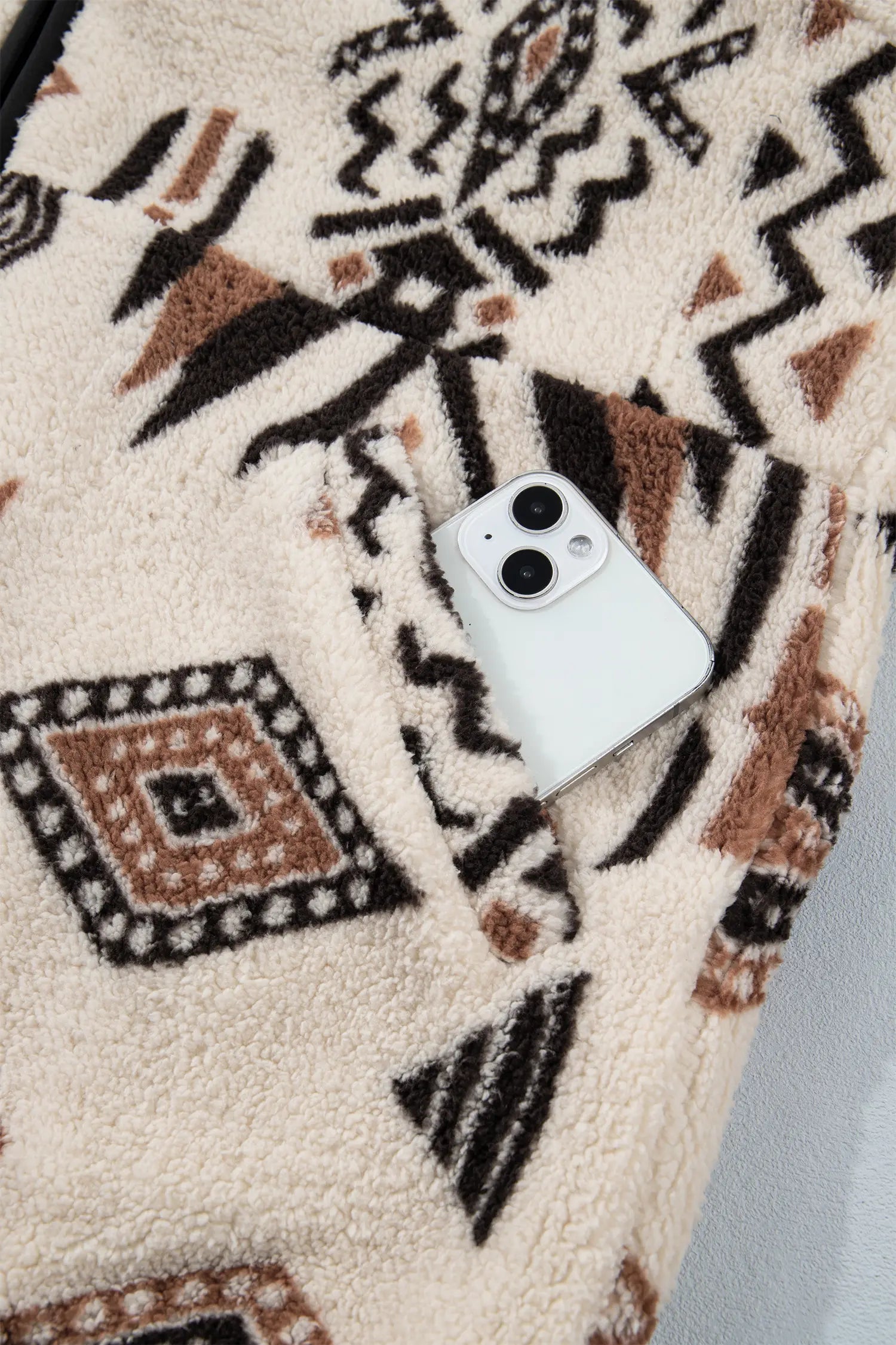 A close-up of a cream sherpa fleece garment with a black and brown geometric pattern, featuring a white smartphone tucked into the pocket.