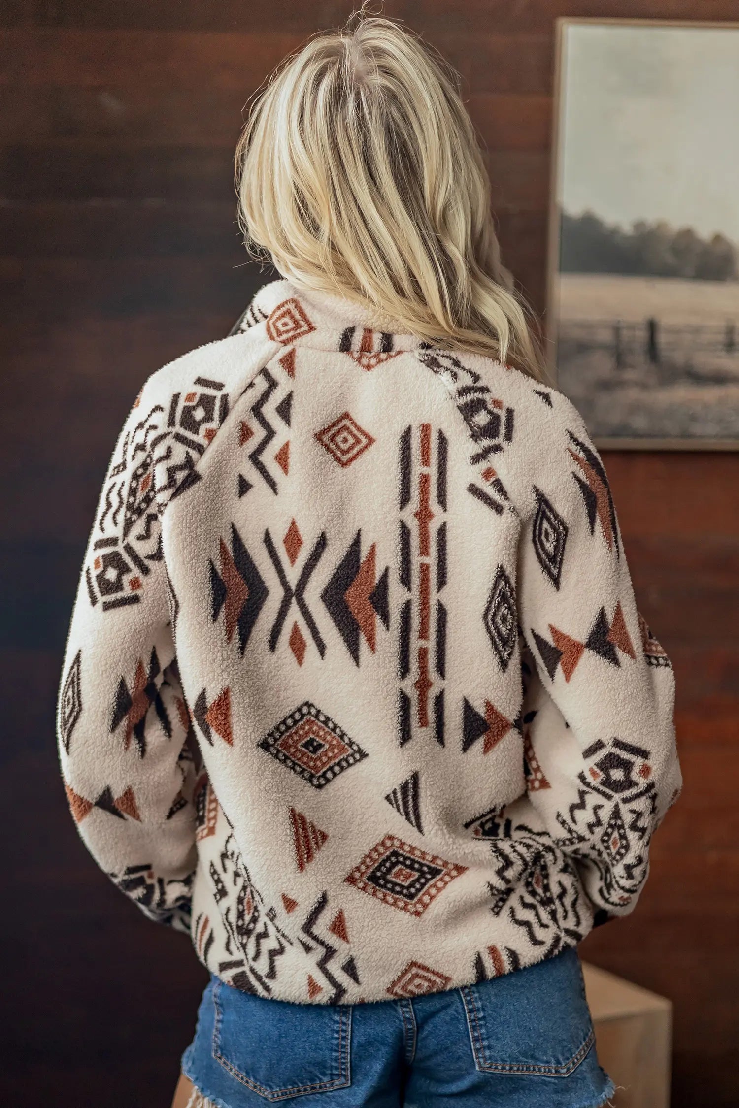 A woman viewed from behind wearing a cream-colored sherpa fleece jacket with a brown and black Southwestern geometric pattern, paired with blue jeans in a rustic indoor setting.