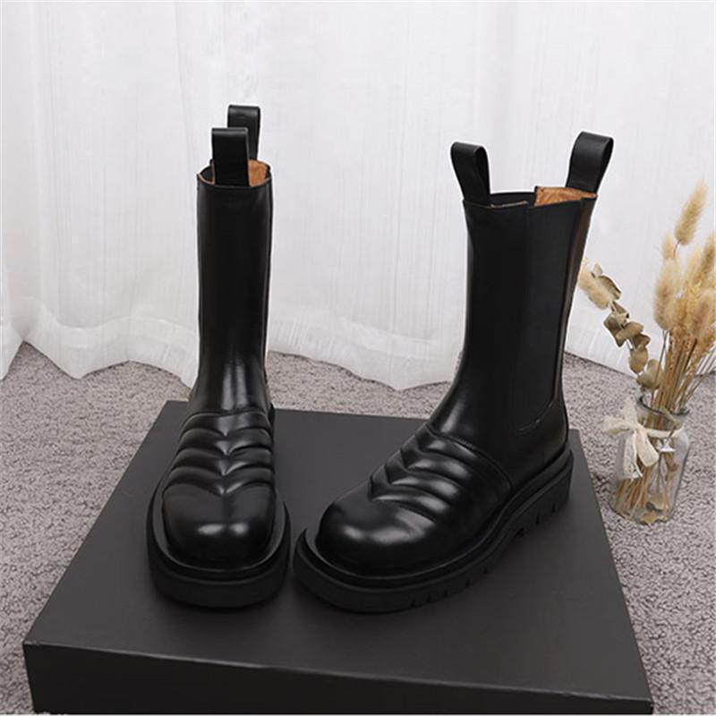 Pair of black leather boots on a black platform with a white curtain and decorative plant in the background.