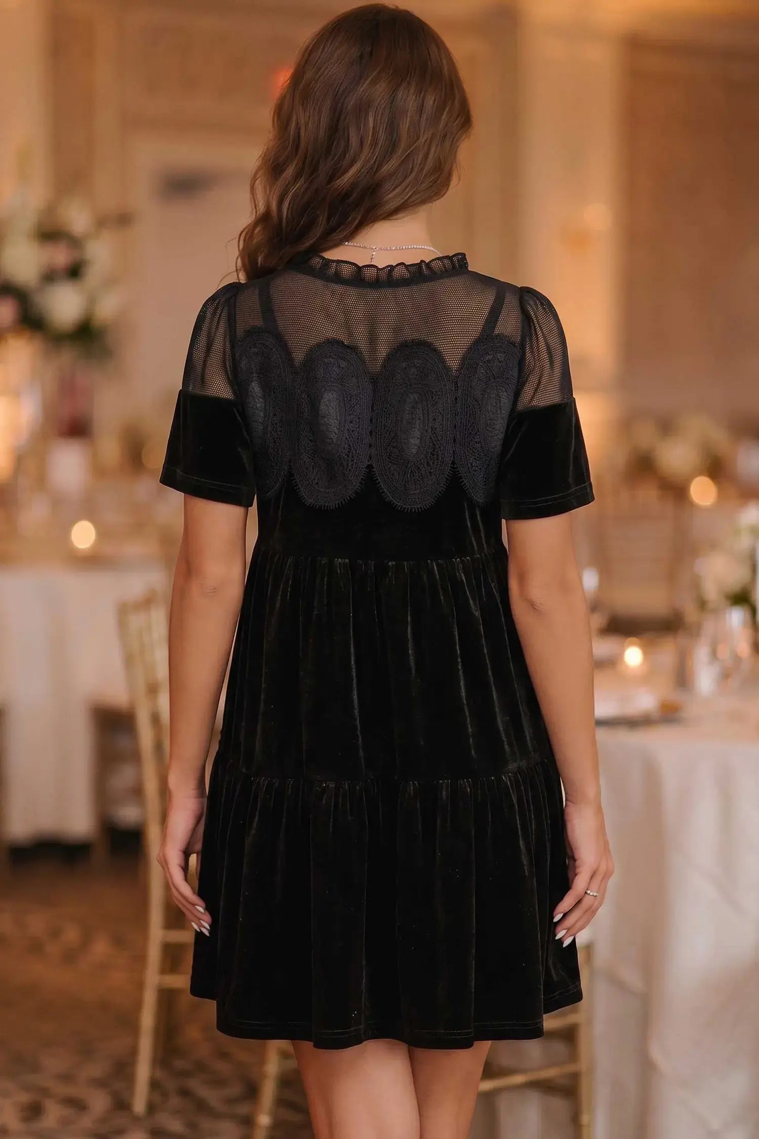 A woman viewed from behind wearing a black velvet dress with sheer mesh back panels and scalloped black lace appliqués, standing in a softly lit event venue with tables and candles in the background.