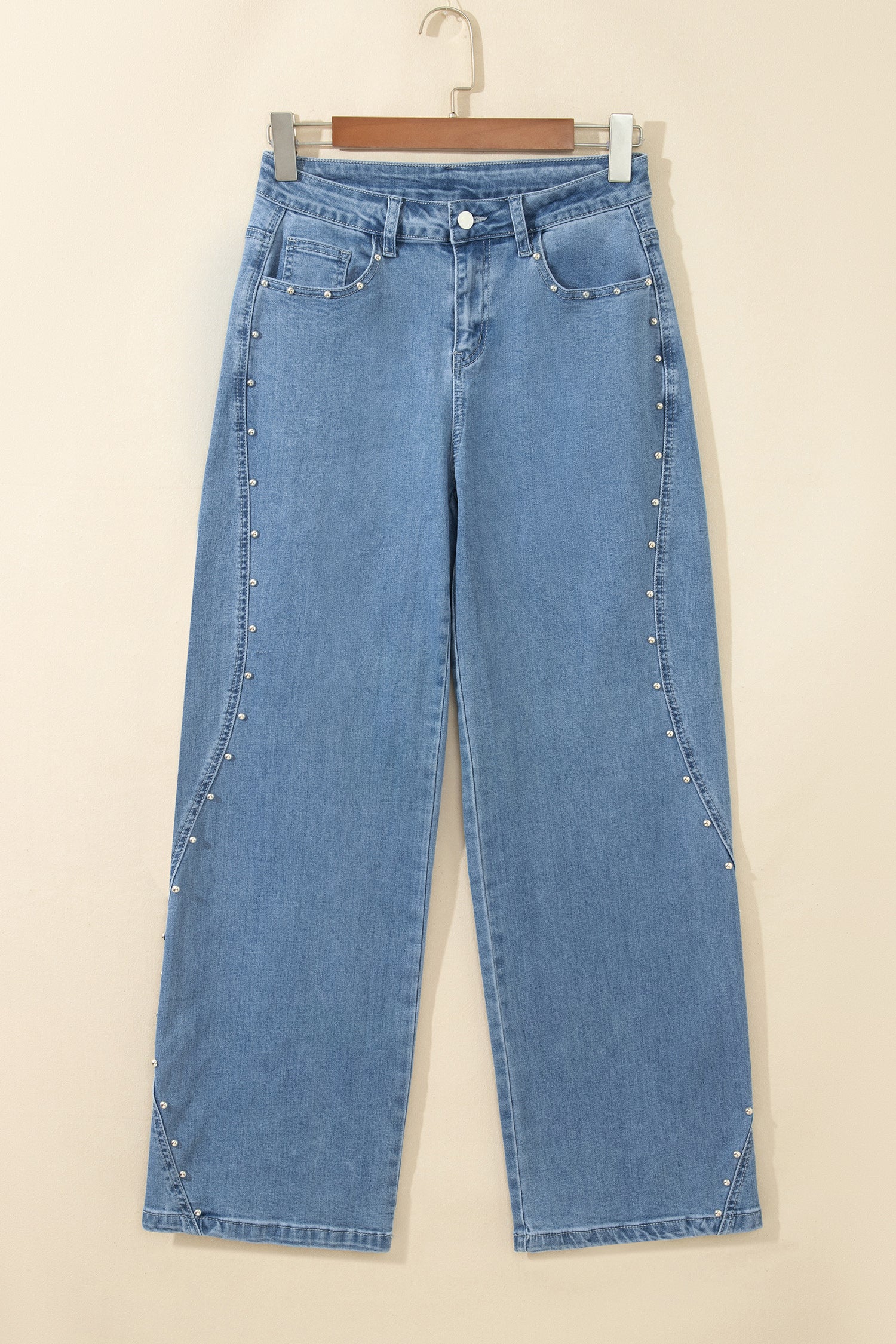 Light blue denim jeans with silver stud embellishments along the side seams, hanging on a wooden hanger against a plain beige wall.