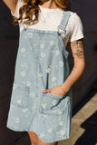 A person wearing light blue denim overall with a white floral print over a white t-shirt, standing outdoors in warm sunlight.