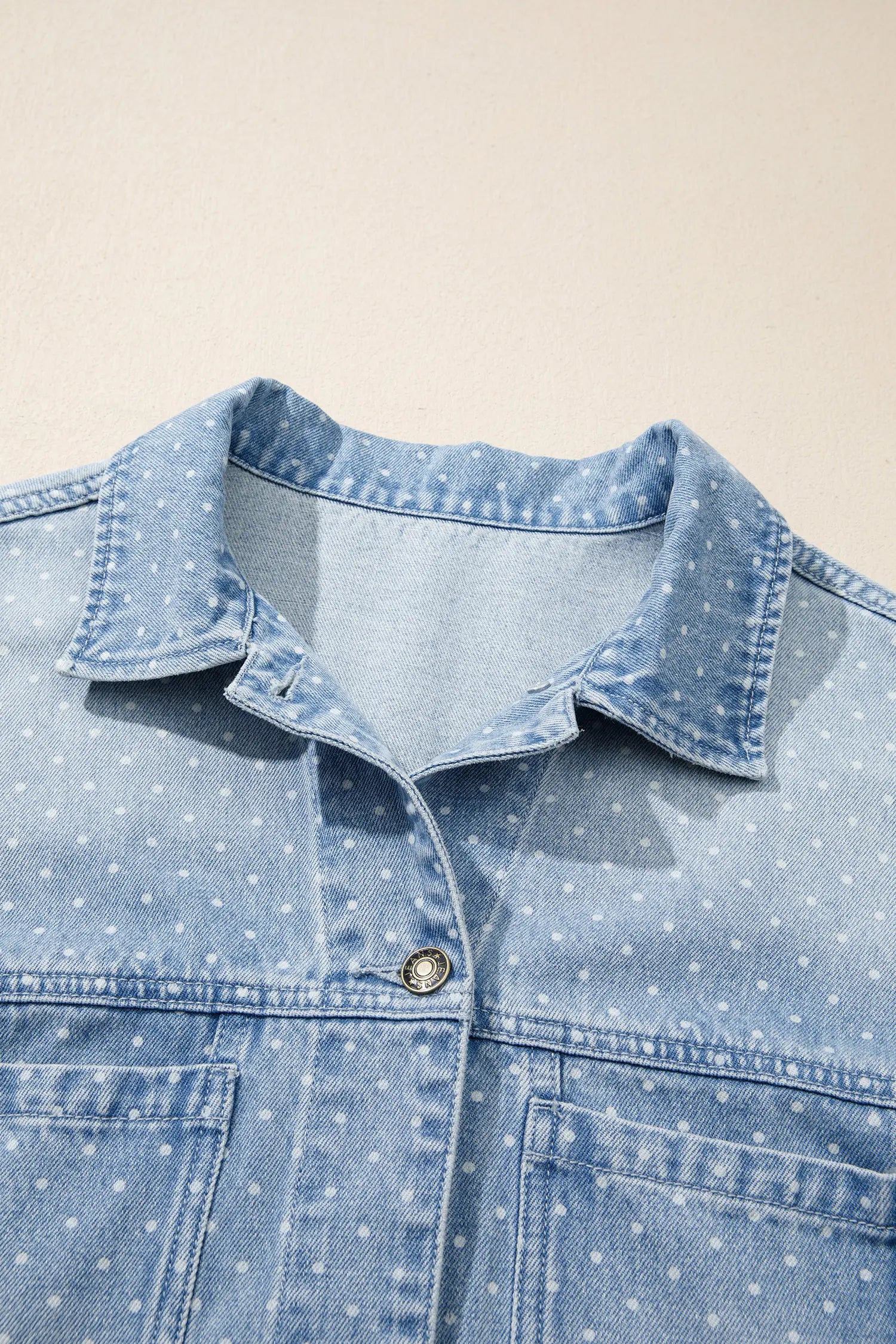 Close-up of light blue denim shirt jacket with white polka dots, pointed collar, and silver snap button.
