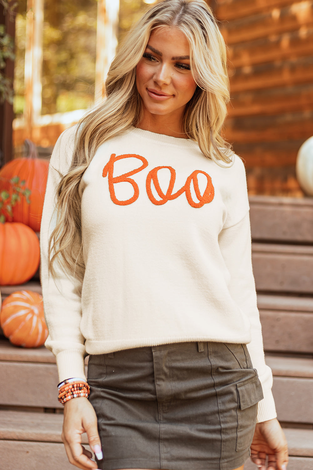 Woman wearing a white sweater with 'Boo' in orange text, standing in front of pumpkins.