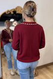 A woman viewed from behind wearing a burgundy long-sleeve knit top with a decorative geometric embroidered collar, paired with light blue jeans. She is standing in front of a mirror in an indoor setting.