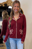 A woman wearing a burgundy zip-up cardigan with beige embroidered trim and bow details on the pockets, paired with light blue jeans. She is standing in front of a mirror in a boutique setting.