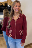 A woman wearing a maroon zip-up cardigan with gold bow and trim details, paired with light blue jeans, standing in front of a mirror.