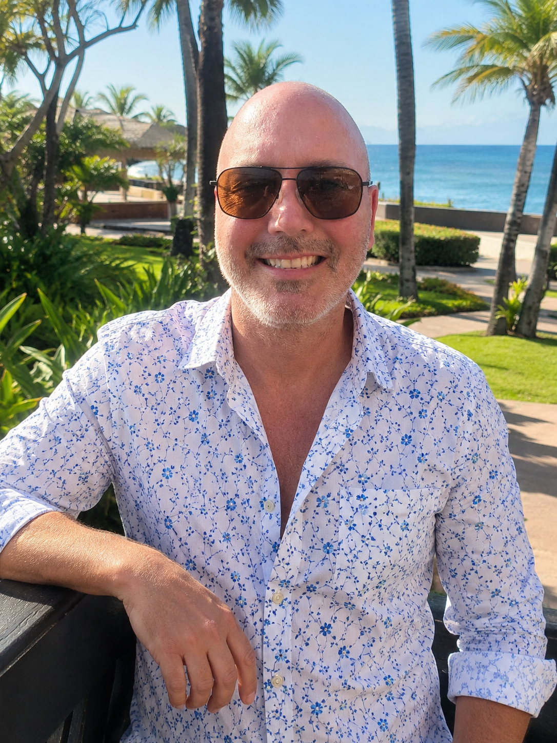 Man wearing sunglasses and a floral shirt in a tropical setting with palm trees and ocean.