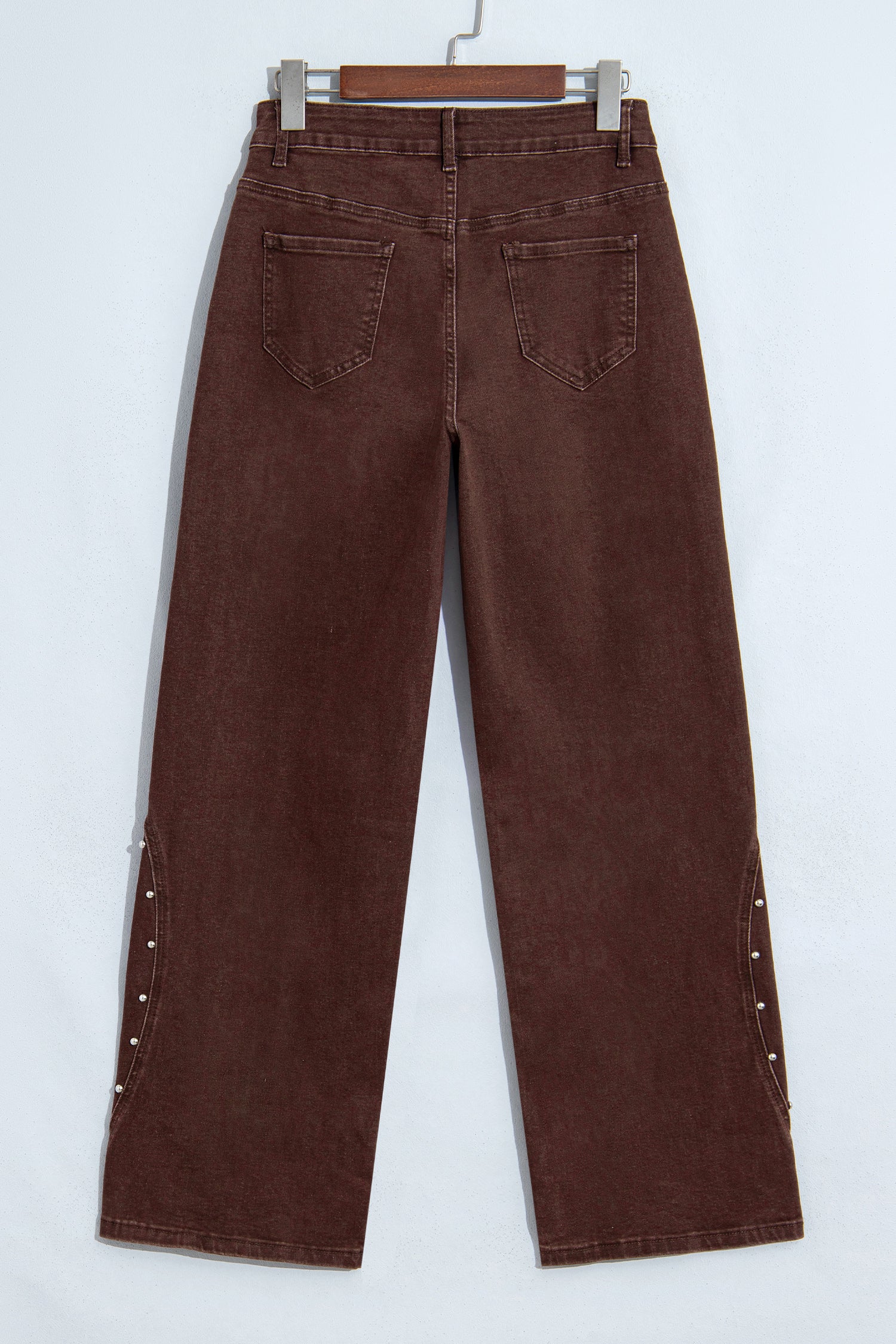 A pair of dark brown wide-leg jeans shown from the back, hanging on a wooden hanger against a plain white wall. The jeans feature decorative silver studs along the outer seams of the legs.
