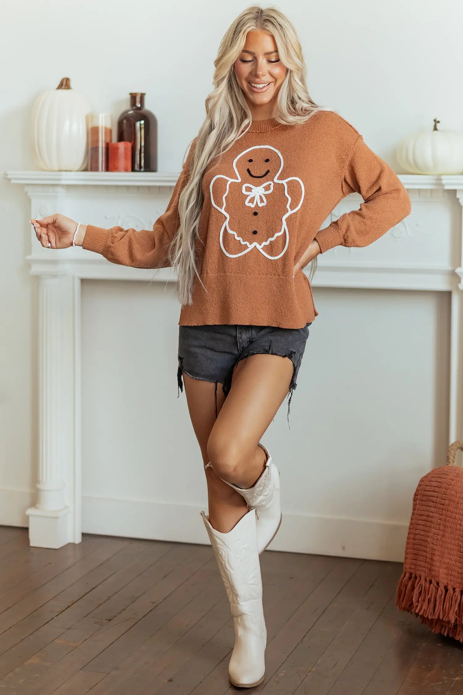 A woman modeling a rust-orange crewneck sweater with a white gingerbread man graphic, paired with denim shorts and white cowboy boots, standing in front of a white fireplace mantel decorated with pumpkins.