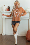 A woman modeling a rust-orange crewneck sweater with a white gingerbread man graphic, paired with denim shorts and white cowboy boots, standing in front of a white fireplace mantel decorated with pumpkins.