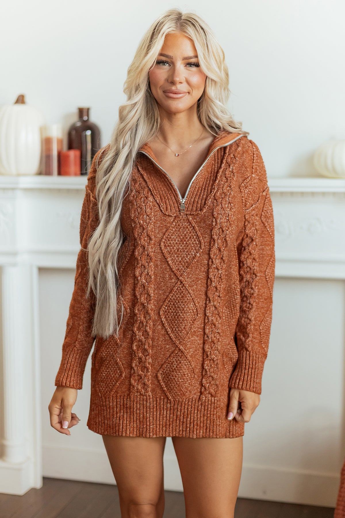 Woman in rust-orange cable-knit half-zip sweater dress standing by white fireplace mantel.