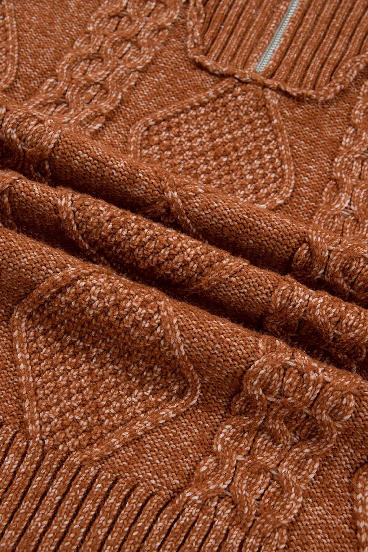 Close-up of a textured knit sweater in a warm rust brown color, showing detailed cable-knit and ribbed patterns.