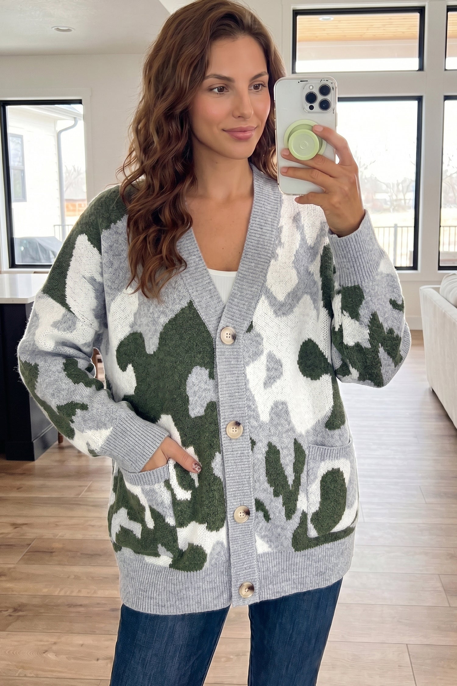A woman takes a mirror selfie wearing a gray knit cardigan with a green and white camouflage pattern, paired with a white top and blue jeans in a bright, modern room.