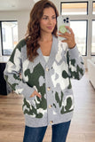 A woman takes a mirror selfie wearing a gray knit cardigan with a green and white camouflage pattern, paired with a white top and blue jeans in a bright, modern room.