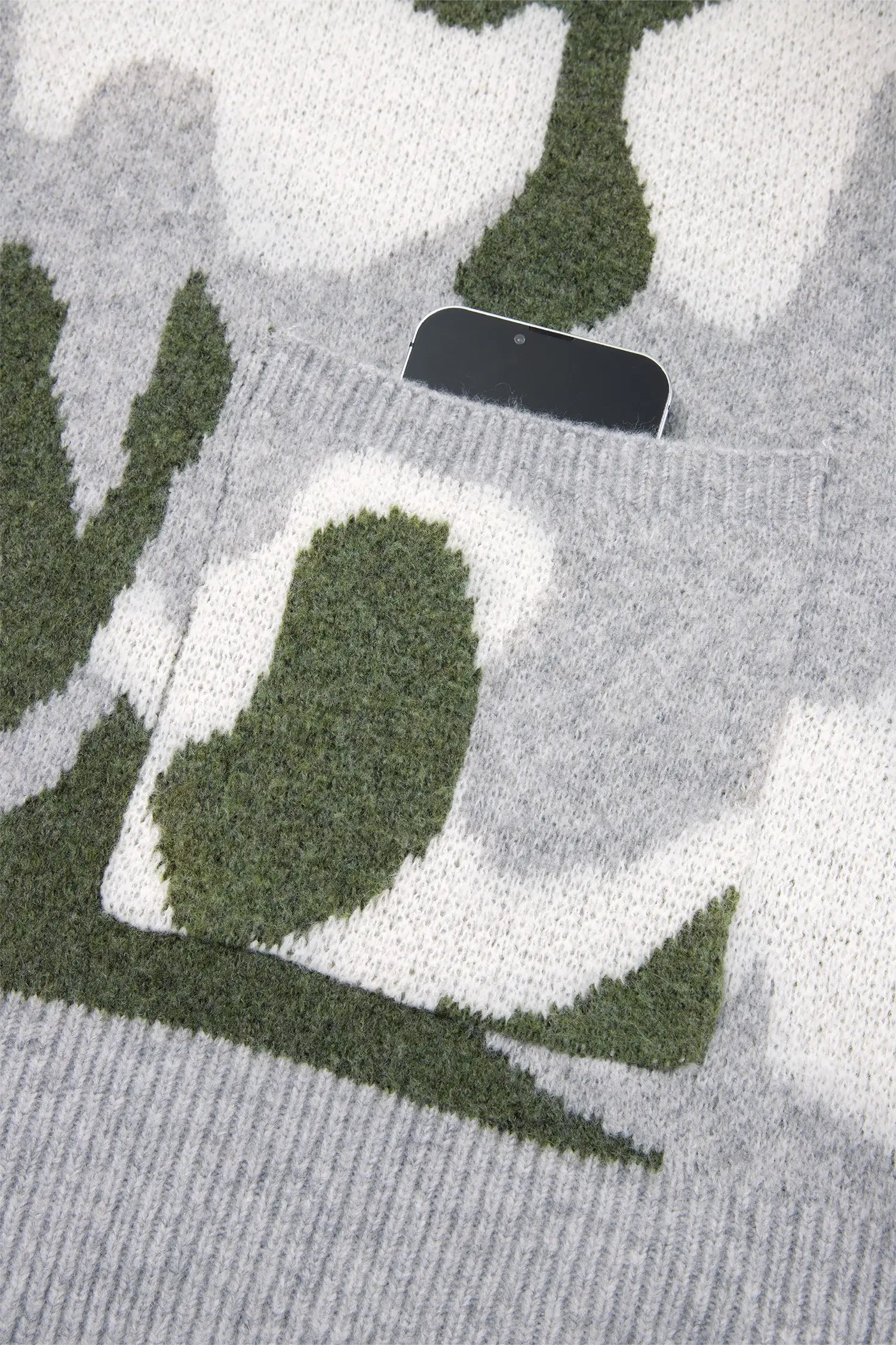 Close-up of a knit sweater with a gray and white camouflage pattern and a green pocket, showing a black smartphone resting inside the pocket.