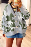 A woman wearing a gray and green camouflage knit cardigan sweater with large buttons, styled with a denim skirt and layered necklaces, in a home setting.