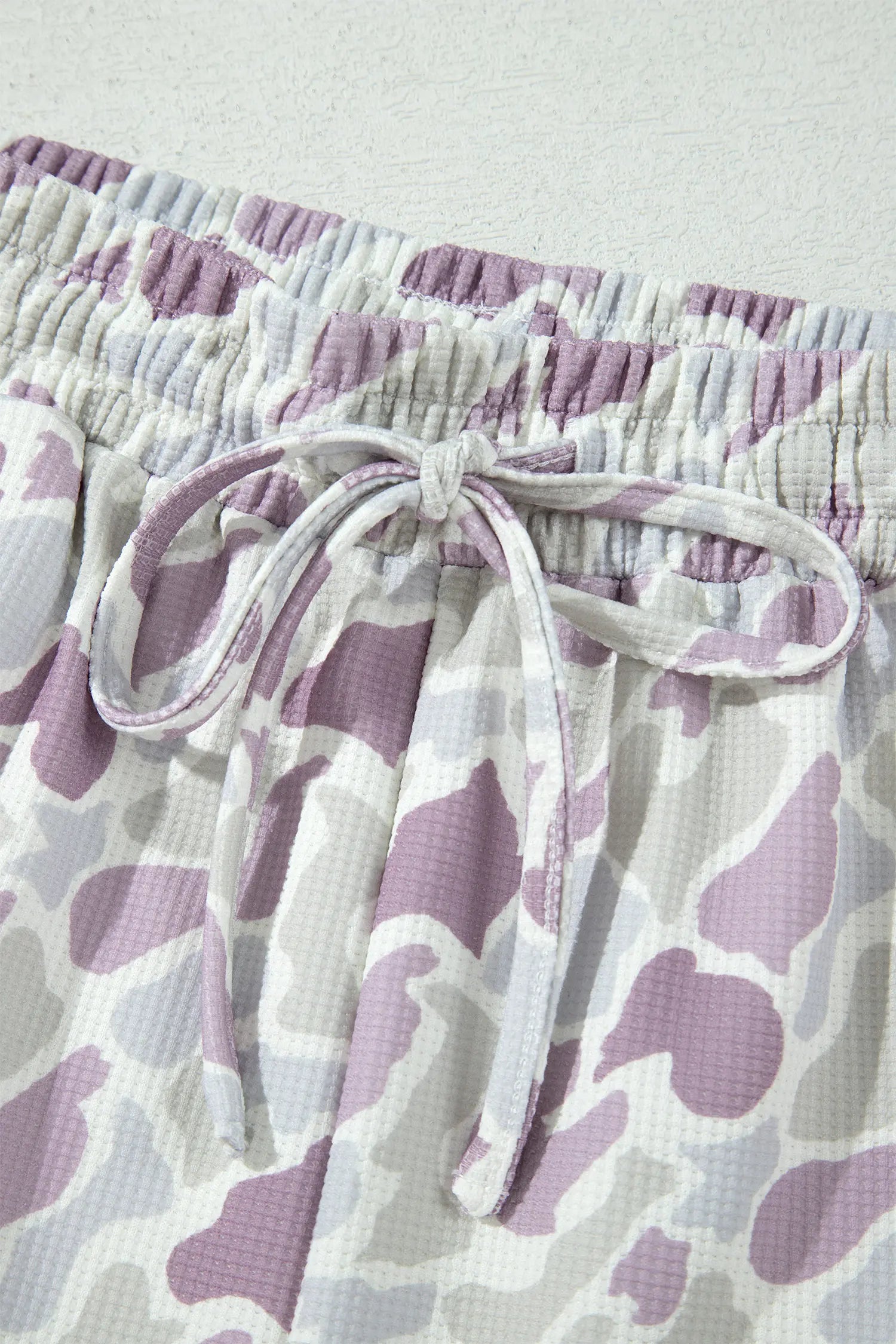 Close-up of a pair of shorts with a purple and gray waffle-textured cow print pattern, featuring an elasticized waistband and matching drawstring tie.