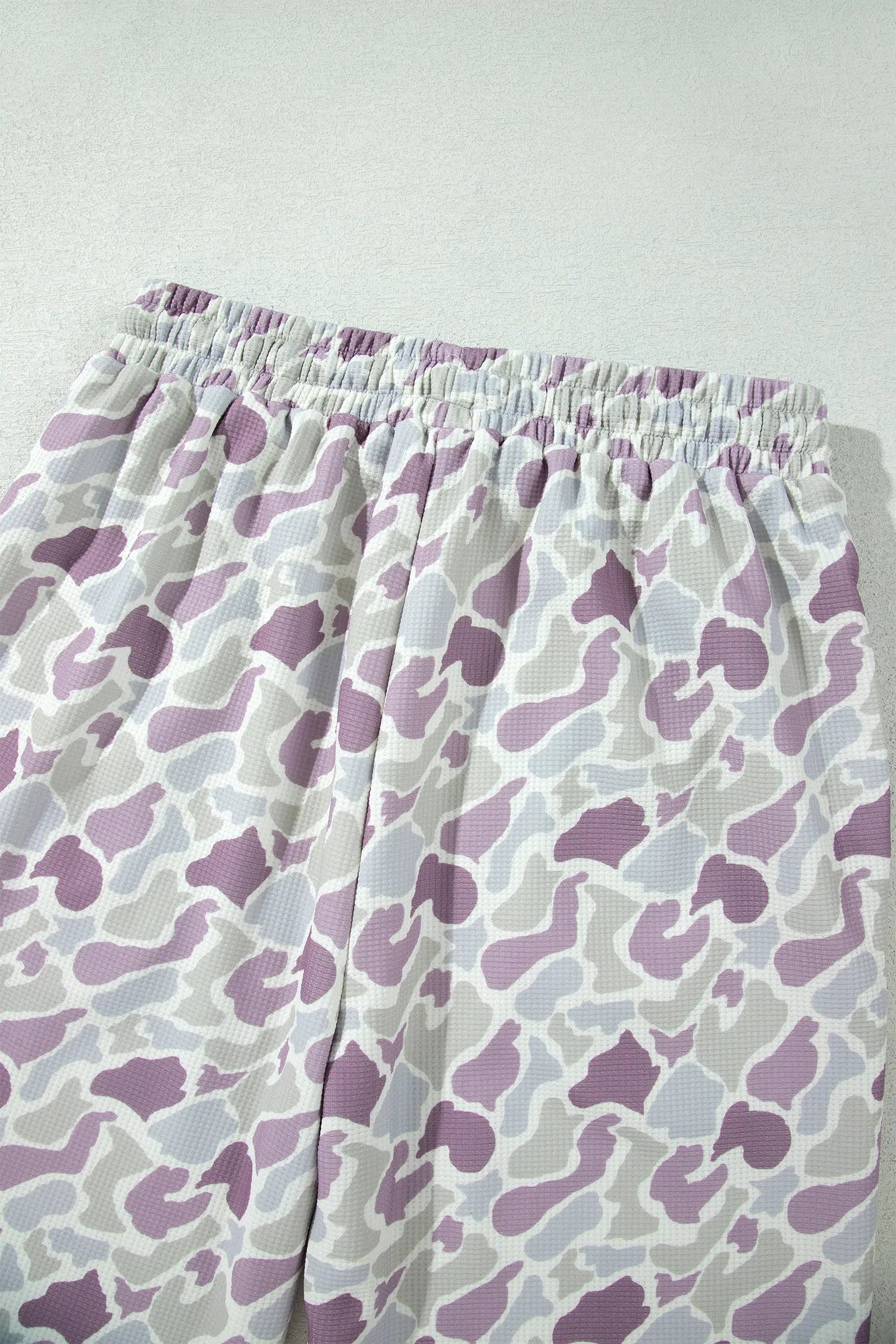 Close-up of a pair of shorts with an elastic waistband, featuring a purple and gray abstract camouflage pattern on a white background.