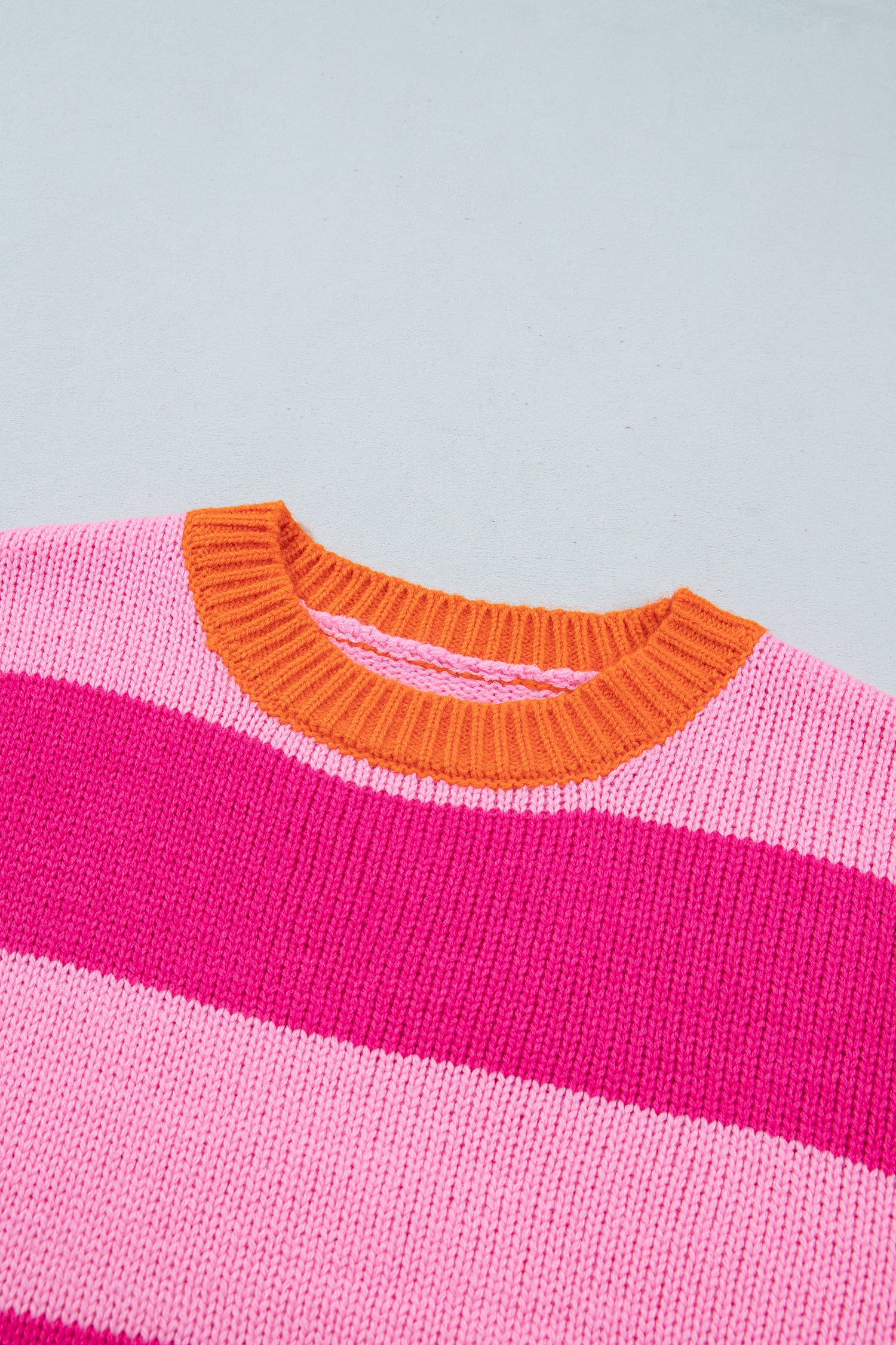 Close-up of a knit sweater with horizontal stripes in light pink, hot pink, and white, featuring a contrasting bright orange ribbed crew neck.