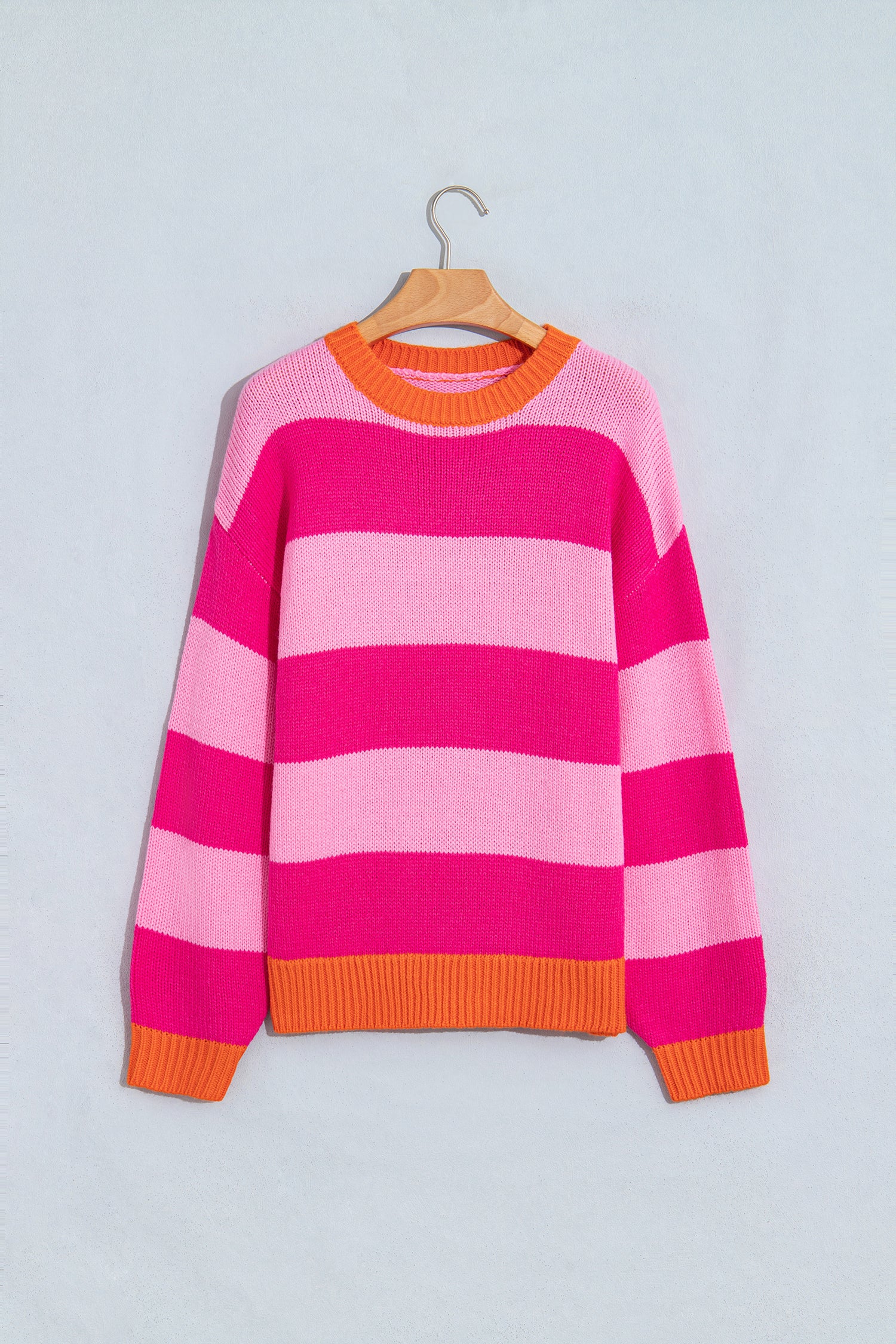 A pink and light pink striped knit sweater with orange ribbed trim, hanging on a wooden hanger against a light gray wall.