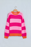A pink and light pink striped knit sweater with orange ribbed trim, hanging on a wooden hanger against a light gray wall.