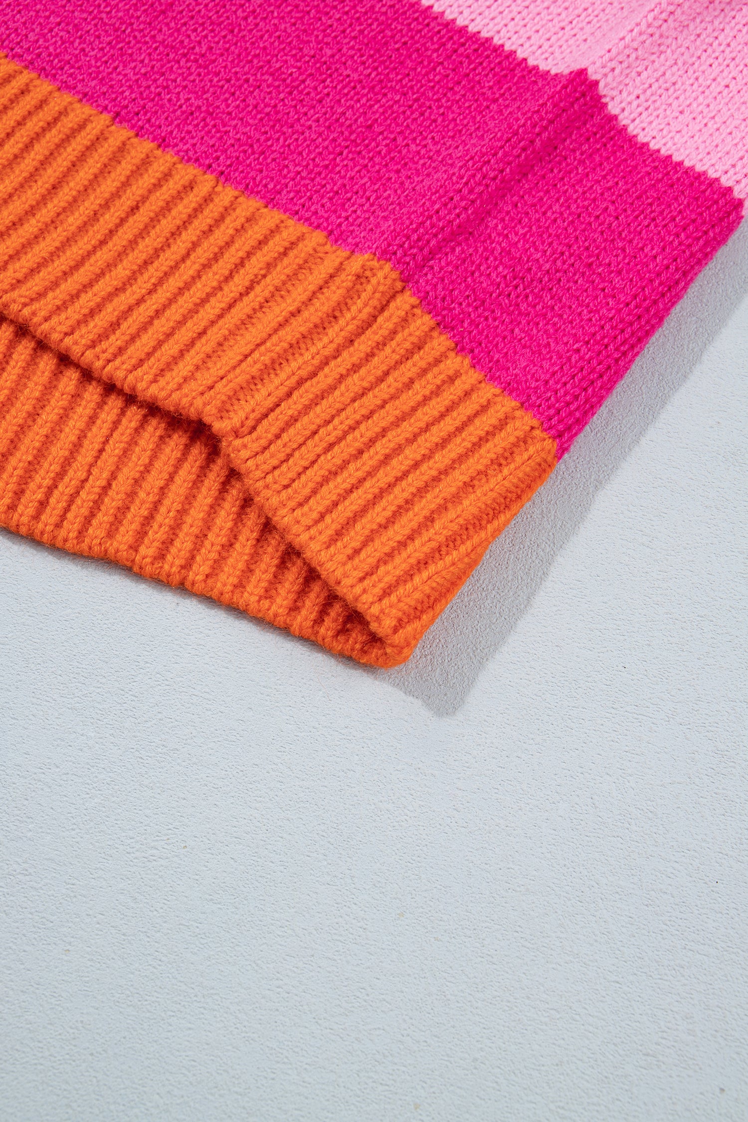 A close-up of a striped knit beanie in bright orange and hot pink, folded diagonally on a light gray textured surface.