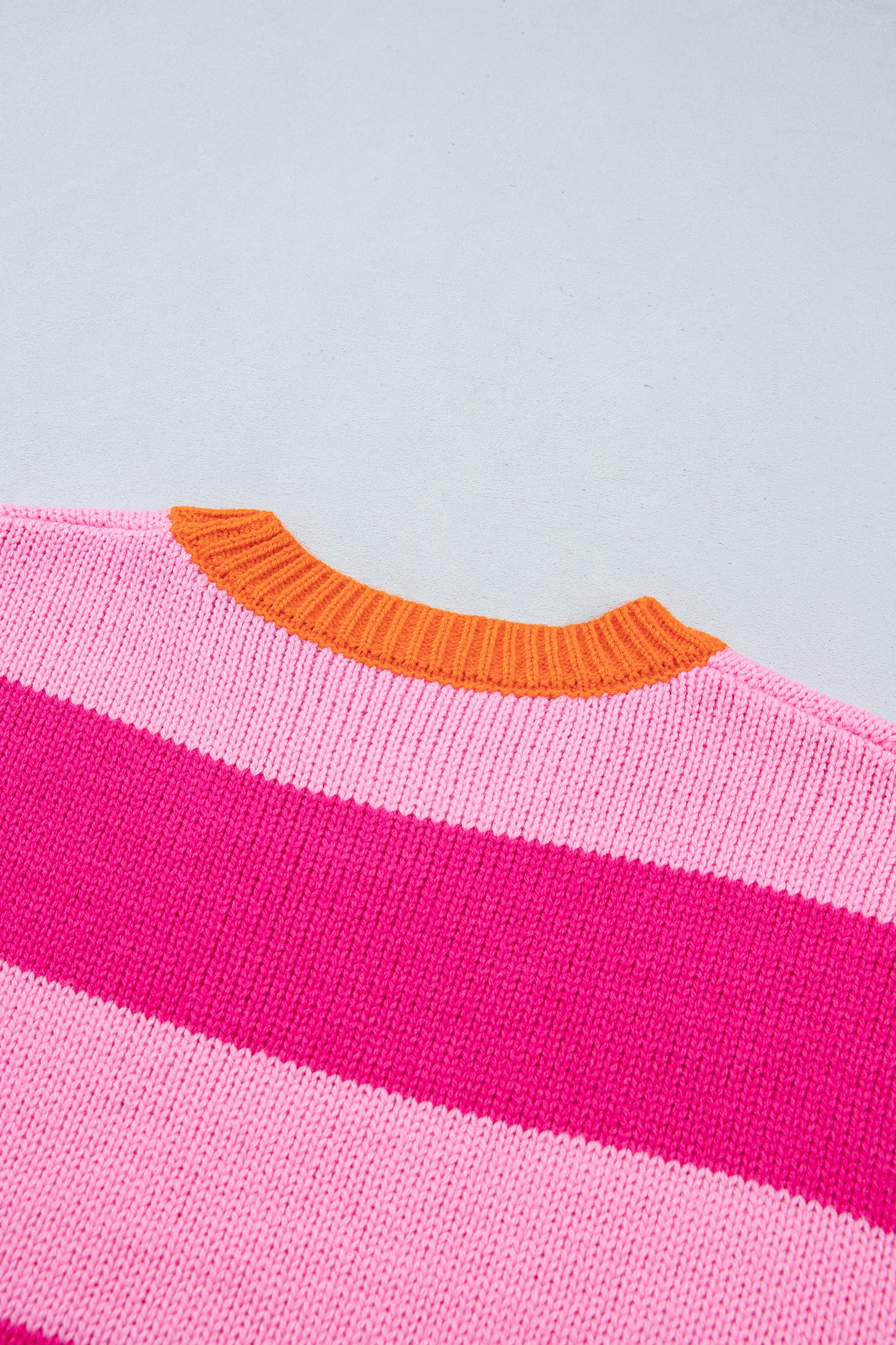 Close-up of a knit sweater showing horizontal pink stripes and a contrasting orange ribbed crew neck, set against a light gray background.