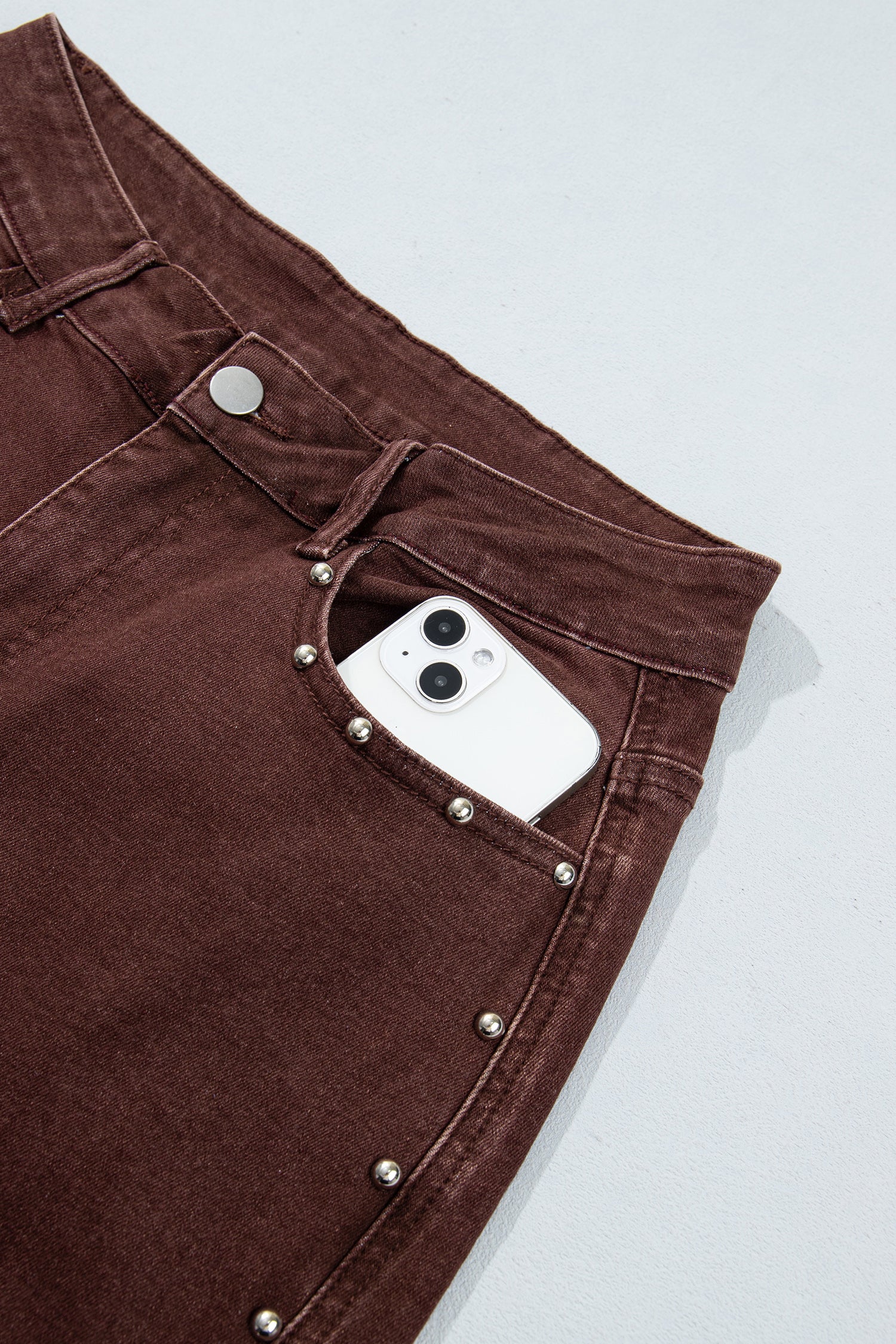 Close-up of dark brown denim jeans with silver stud detailing along the pocket edge, shown with a white smartphone tucked into the back pocket on a light gray background.