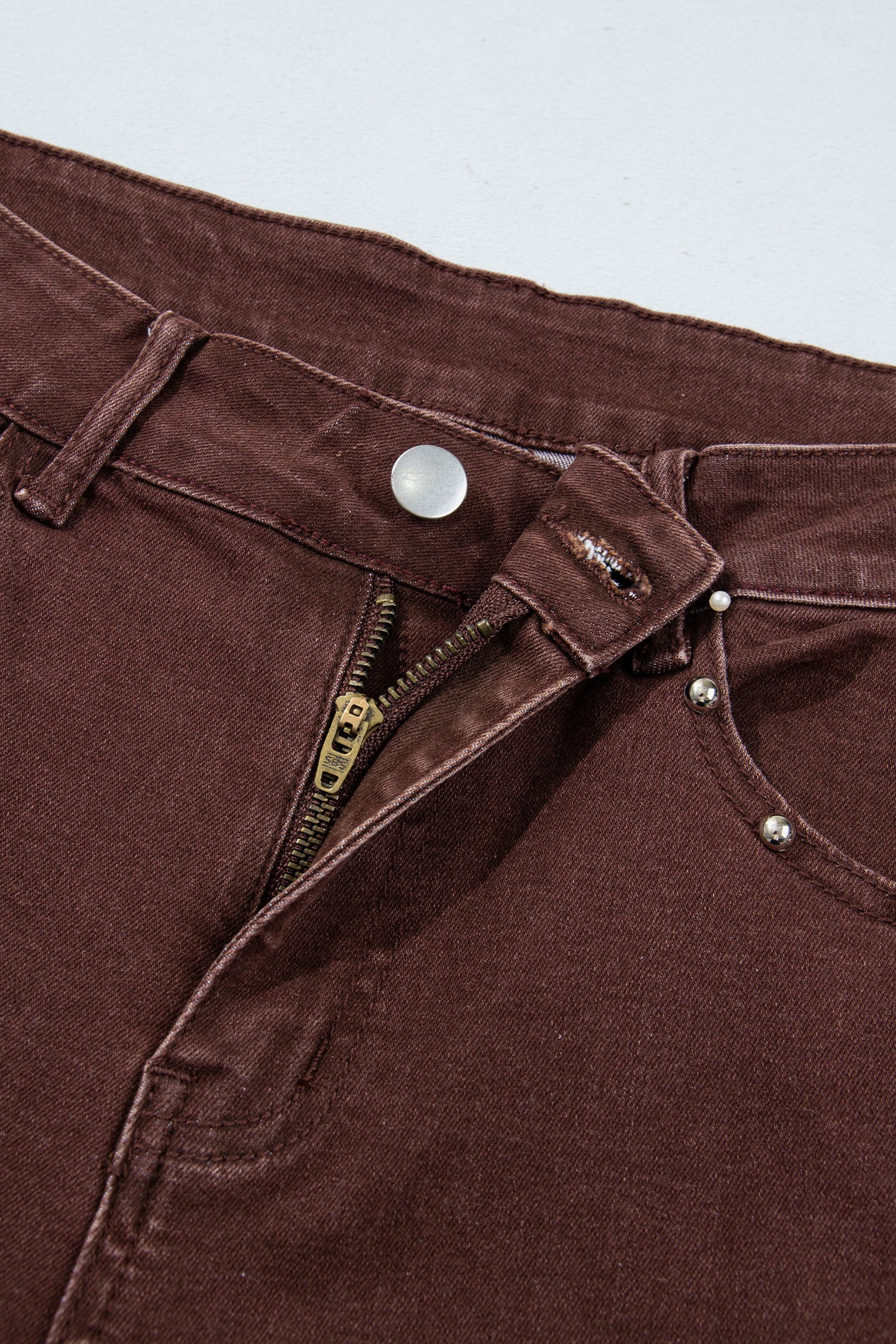 Close-up of dark brown denim pants showing the waistband, button, zipper, rivets, and pocket details against a light gray background.