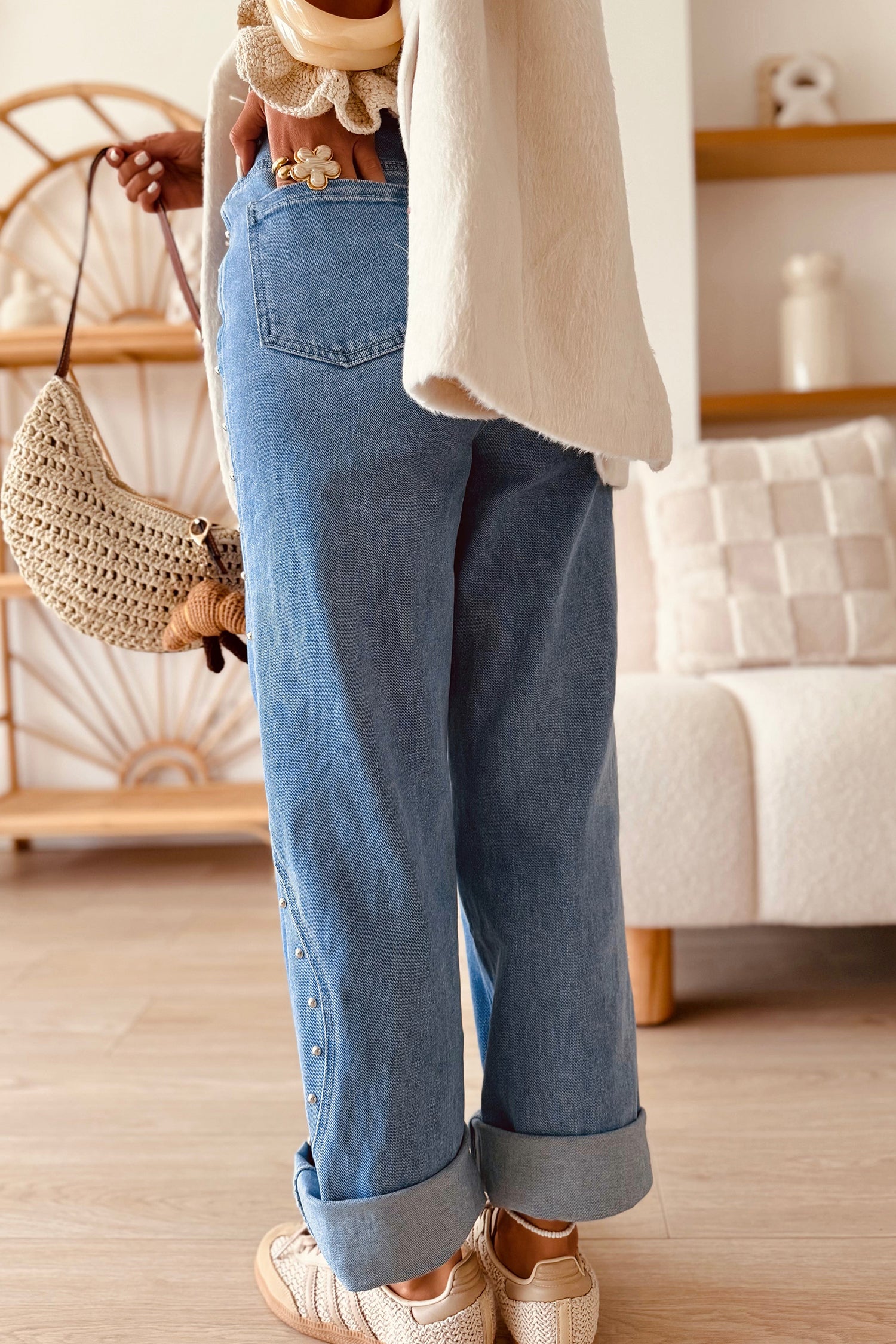 A person wearing light blue wide-leg denim jeans with rolled cuffs, styled with a cream cardigan, beige sneakers, and a woven straw bag in a cozy indoor setting.