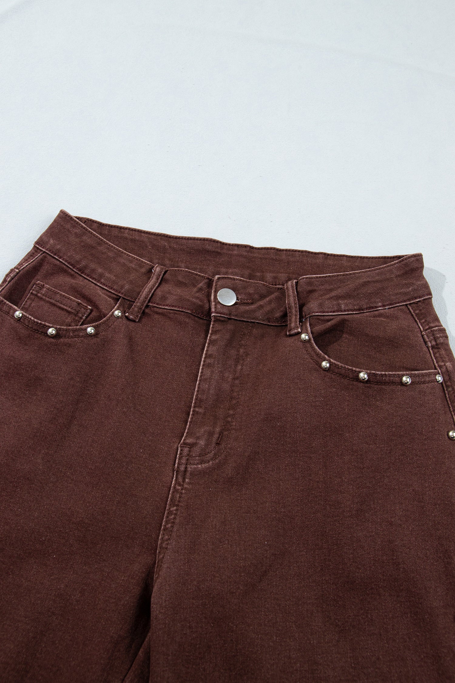 Close-up of dark brown denim jeans showing the waistband, belt loops, button, front pockets, and decorative silver rivets on a light gray background.
